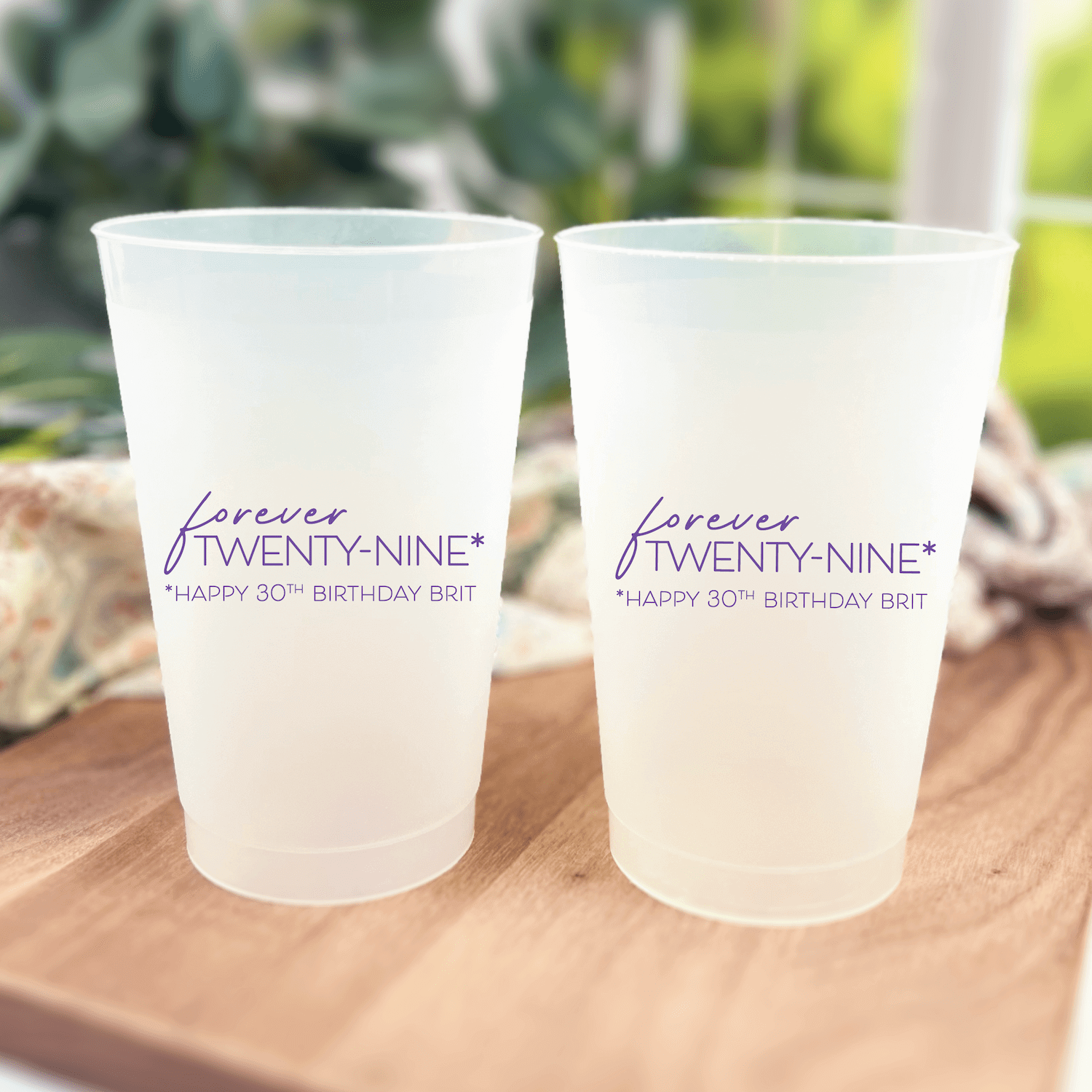 Two frosted plastic cups with "Forever Twenty-Nine Happy 30th Birthday Bash" text printed in navy script, displayed on a wooden surface with greenery background.
