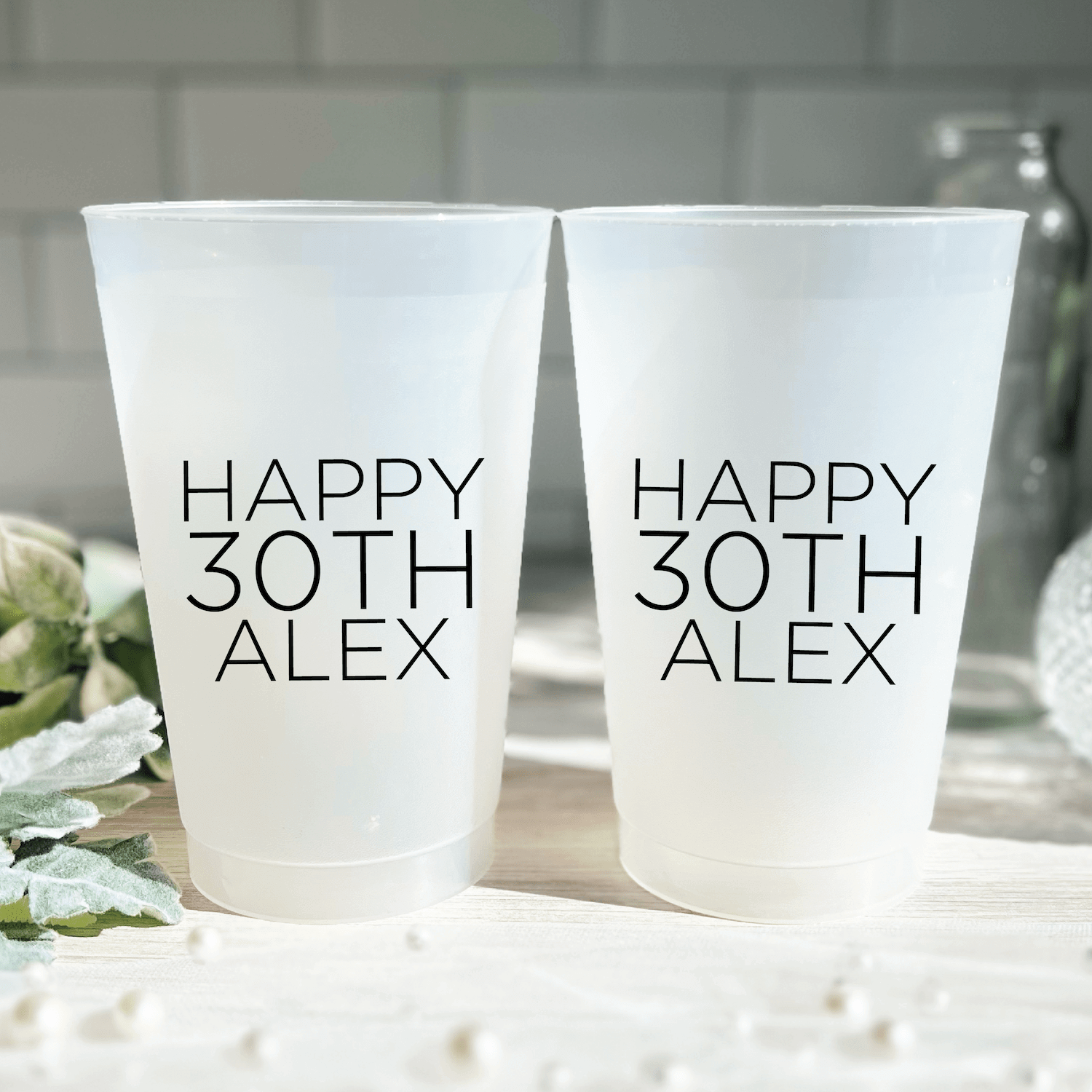 Two frosted plastic cups with "Happy 30th Alex" printed in modern text, displayed on a white surface with white flowers.
