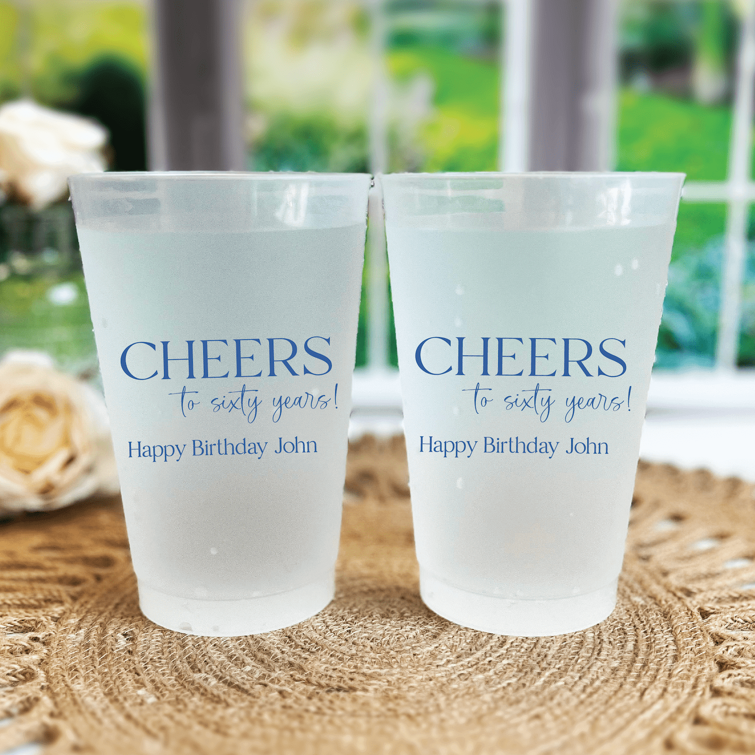 Two frosted plastic cups printed with "Cheers to sixty years" and "Happy Birthday John" in blue text, displayed on a woven placemat.