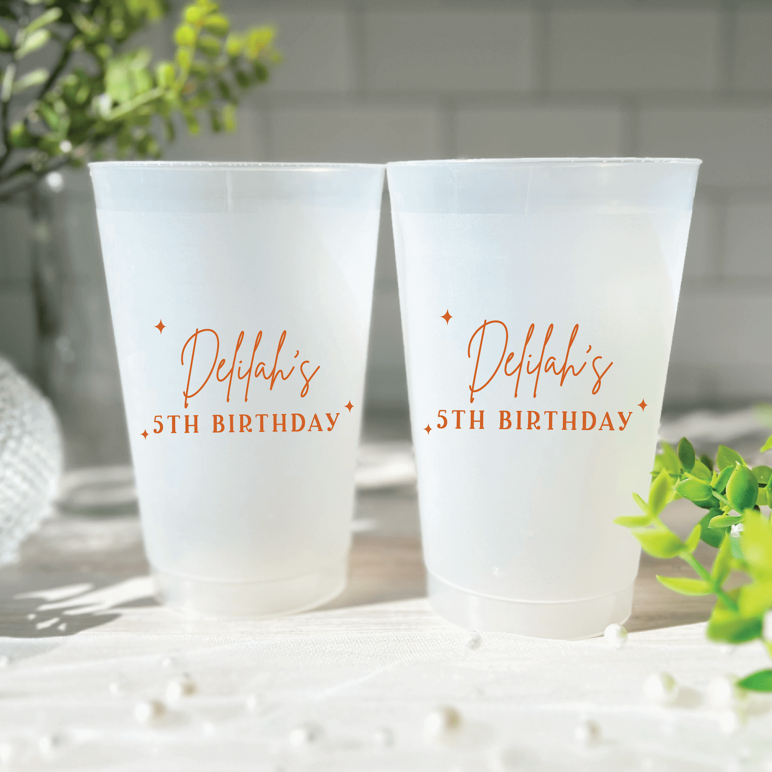 Two frosted plastic cups with orange text reading "Debby's 35th Birthday" with decorative stars, displayed outdoors with greenery.