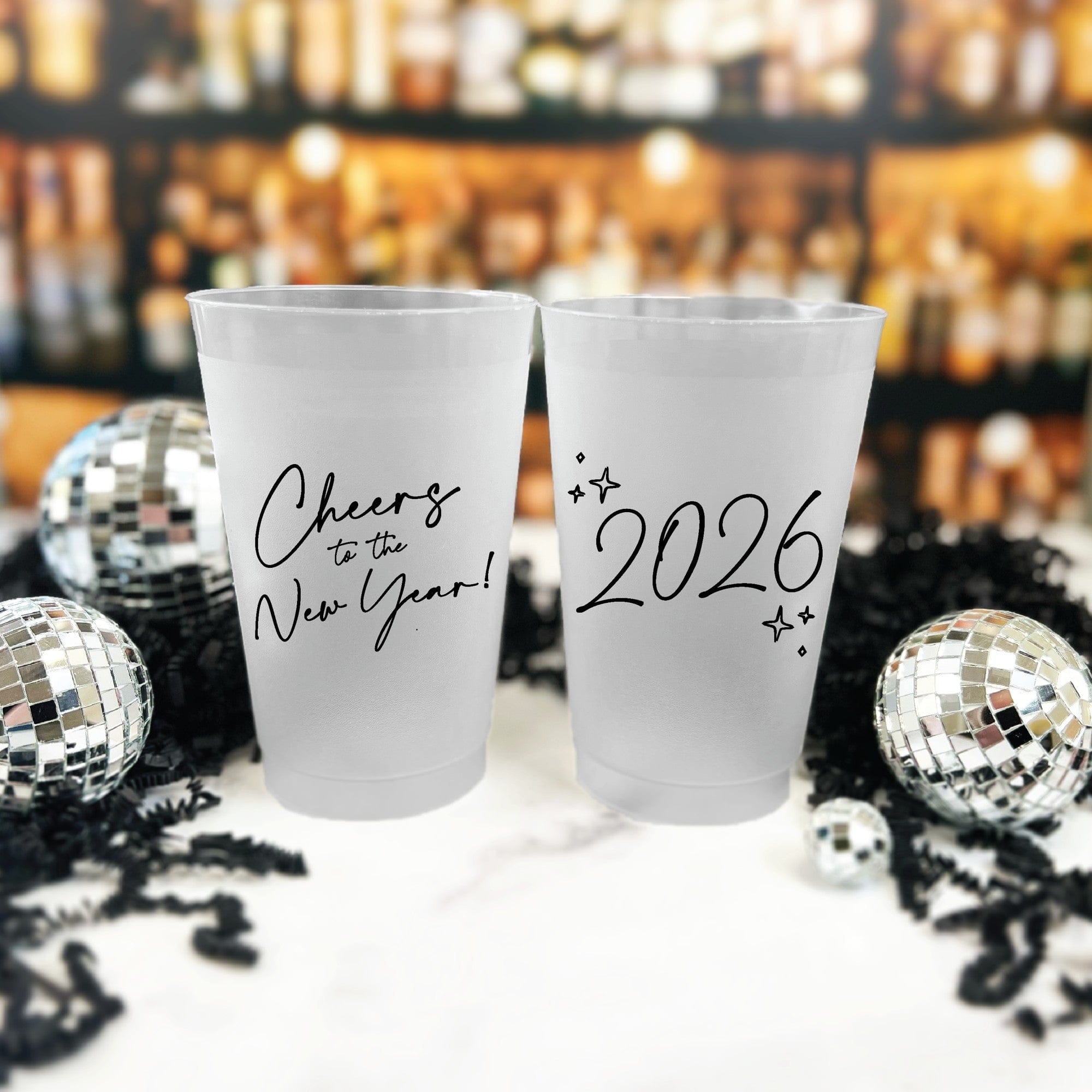 Two frosted plastic party cups are displayed with reflective disco ball decorations and black shredded paper against a blurred bar background, perfect for a New Year's celebration.