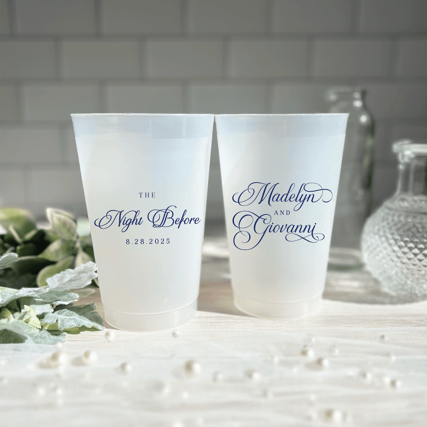 Frosted plastic cups with navy script text reading "The Night Before" and "Madelyn's Rehearsal" with dates, displayed on white surface with flowers and pearls