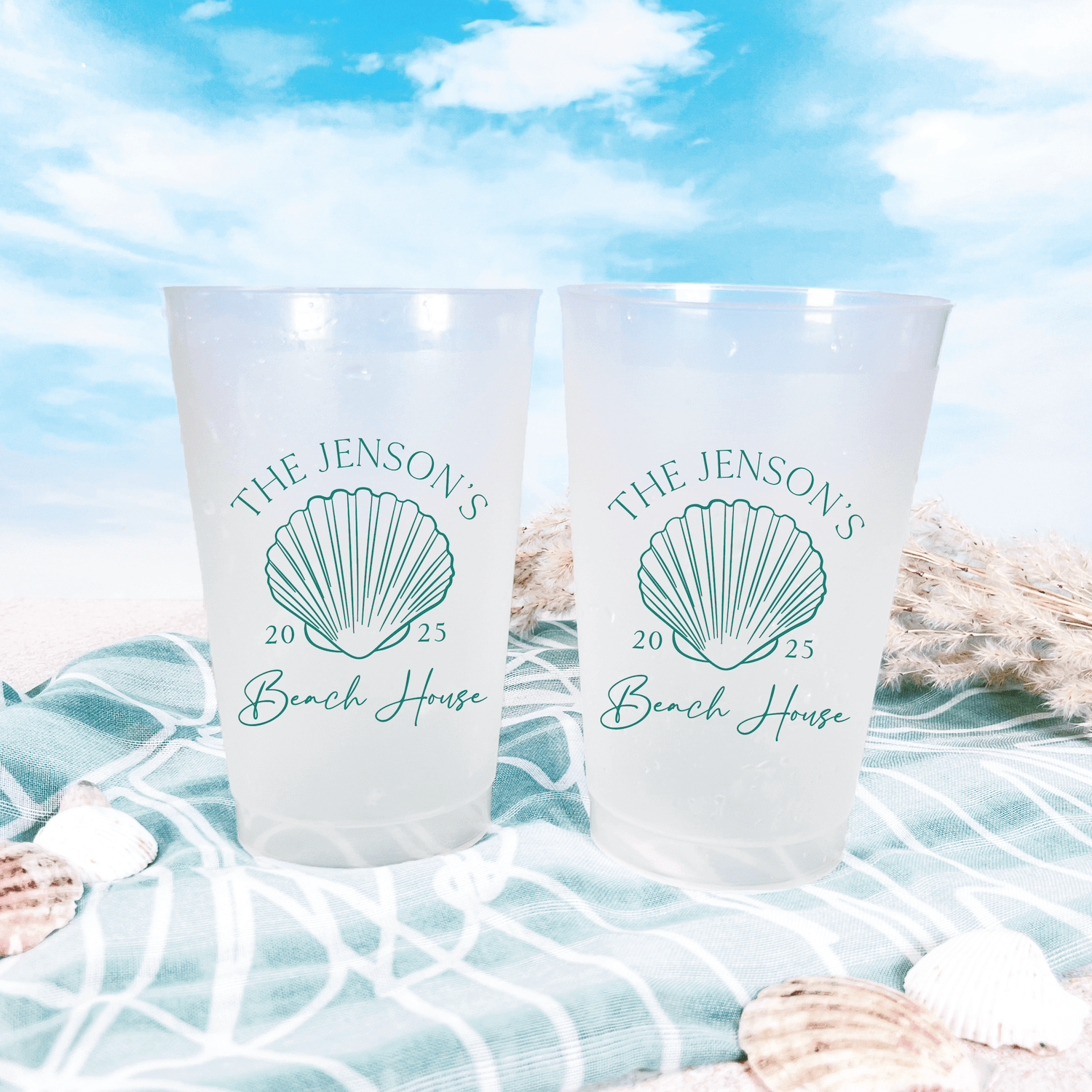 Pair of frosted shatterproof cups with teal seashell design and "The Jensons 2025 Beach House" personalization text, displayed on beach-themed fabric with shells and water backdrop.