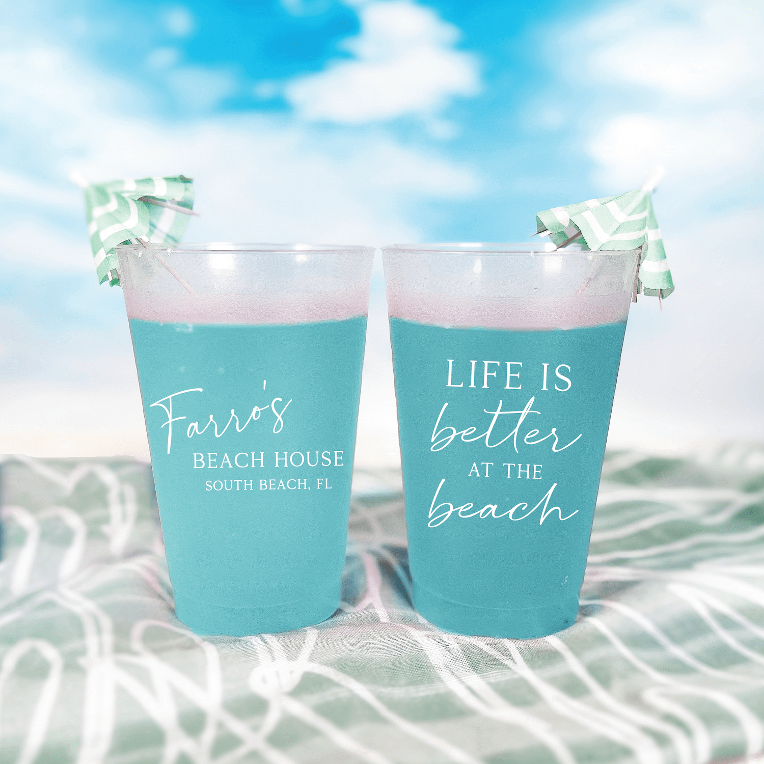 Two turquoise frosted cups with "Life is Better at the Beach" text and personalized beach house details, displayed in shallow water with decorative bow ties.
