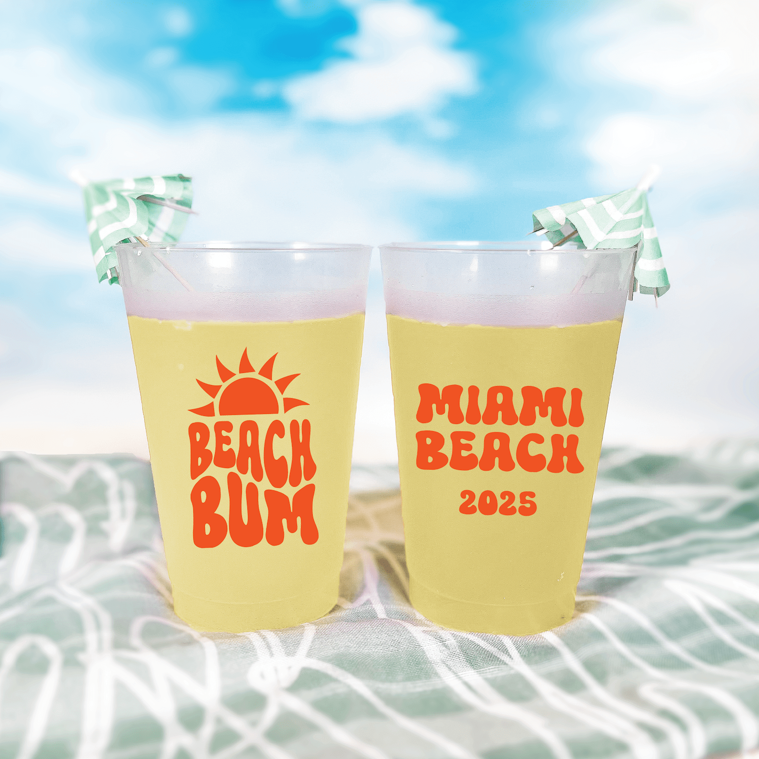 Two yellow frosted cups with "Beach Bum" and "Miami Beach 2025" in red text, topped with green palm leaf decorations, on beach towel background.