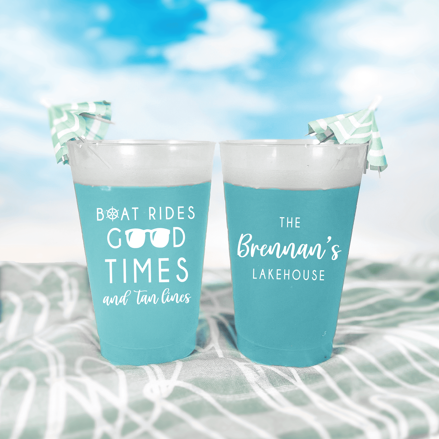 Two turquoise frosted cups with "Boat Rides Good Times and Tan Lines" and "The Bremann's Lakehouse" text, displayed in water with sky background.