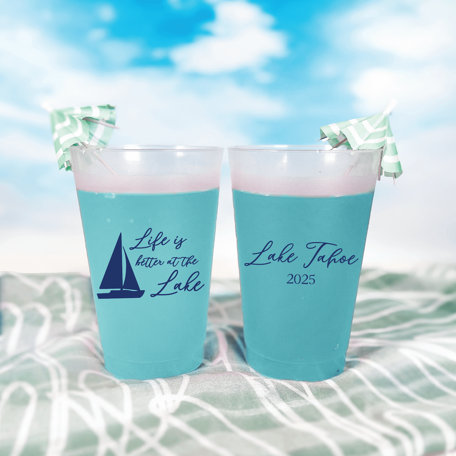 Turquoise frosted cups with sailboat design and "Life is Better at the Lake" text, personalized with "Lake Tahoe 2025," displayed outdoors by water.