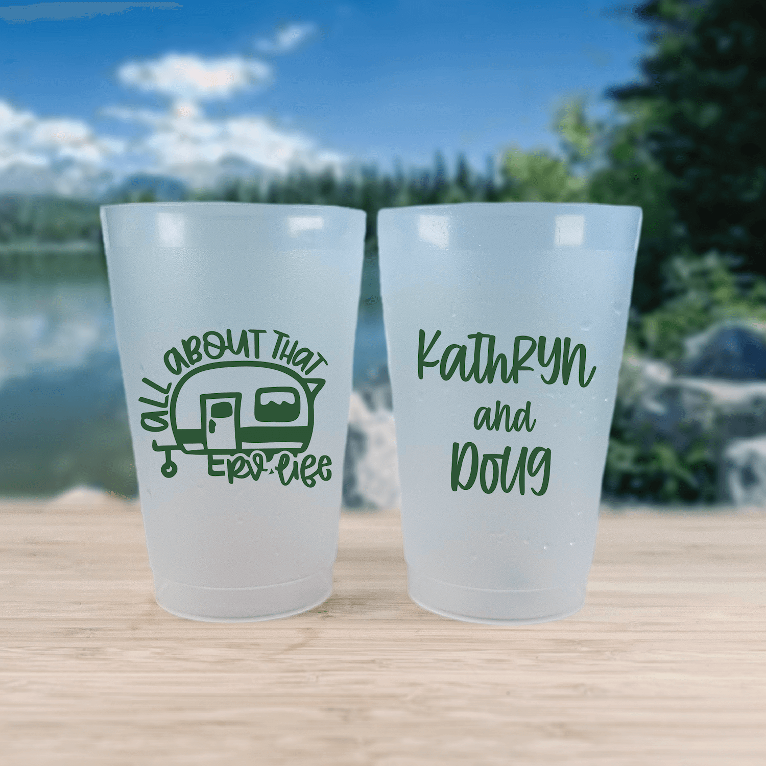 Two frosted cups with green text designs by a lake, one featuring a camper trailer with "All About That RV Life" and the other personalized with "Kathryn and Doug"