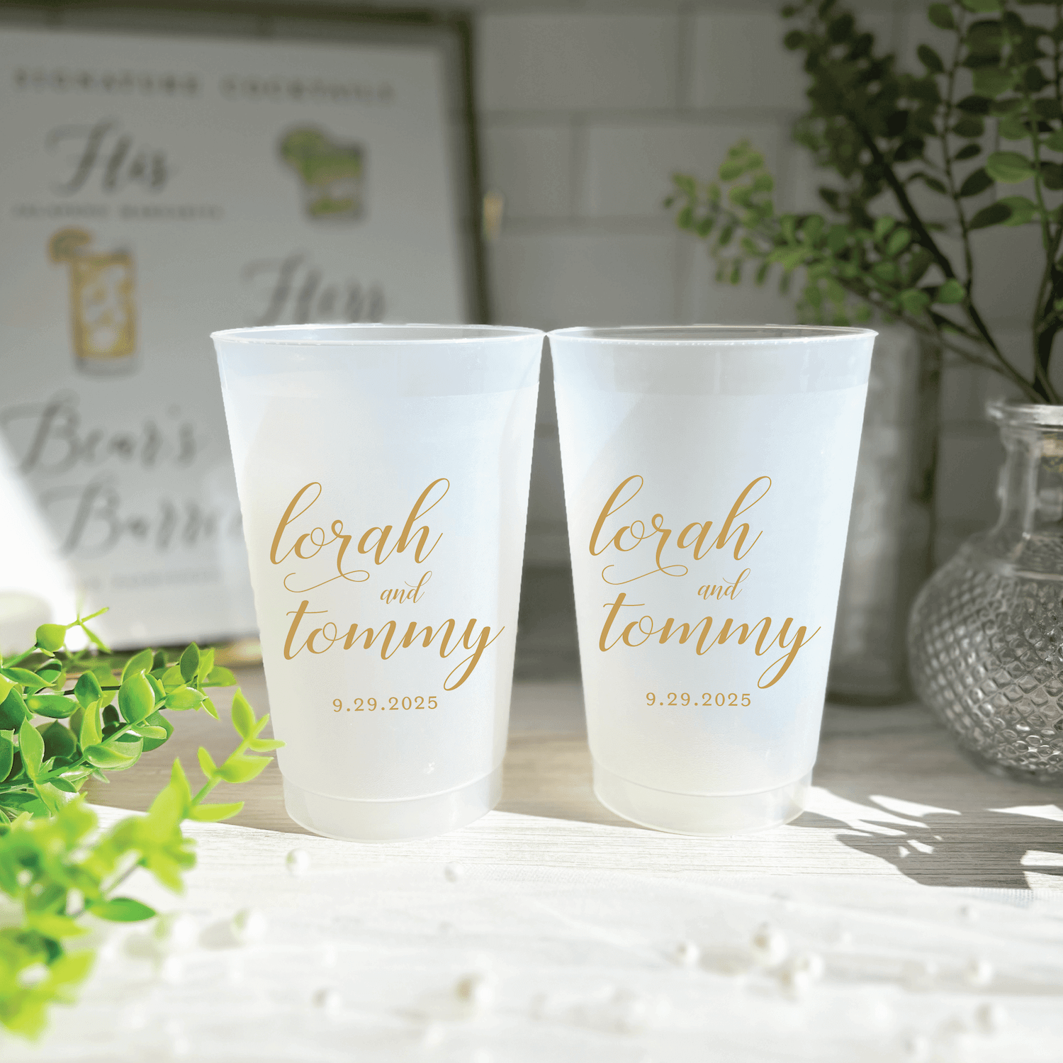 Two frosted plastic cups with gold script reading "lorah and tommy" and "9.25.2023" personalized for a wedding.