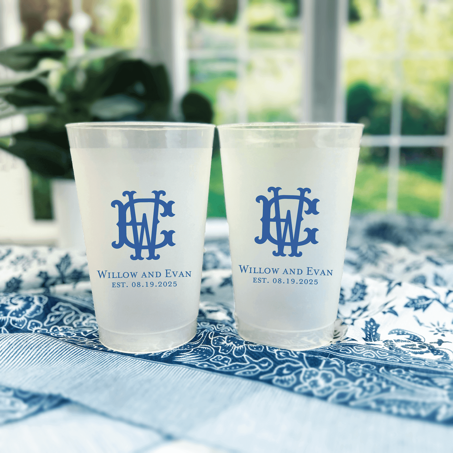 Two frosted cups with blue interlocking monogram and "Willow and Evan EST. 05.17.2025" text on white surface with blue patterned napkins