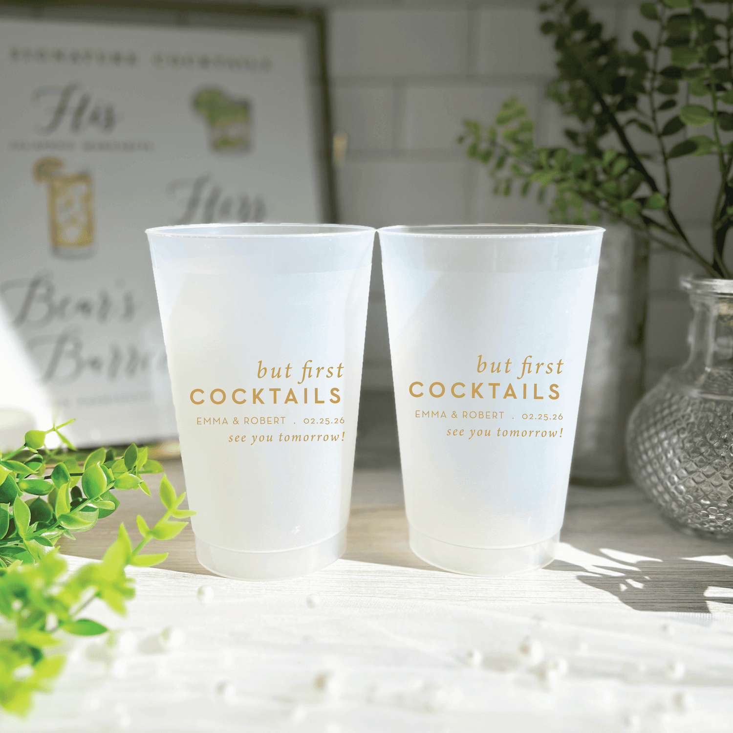 Two frosted cups with gold "but first COCKTAILS" text and personalization details, displayed on a white surface with greenery.
