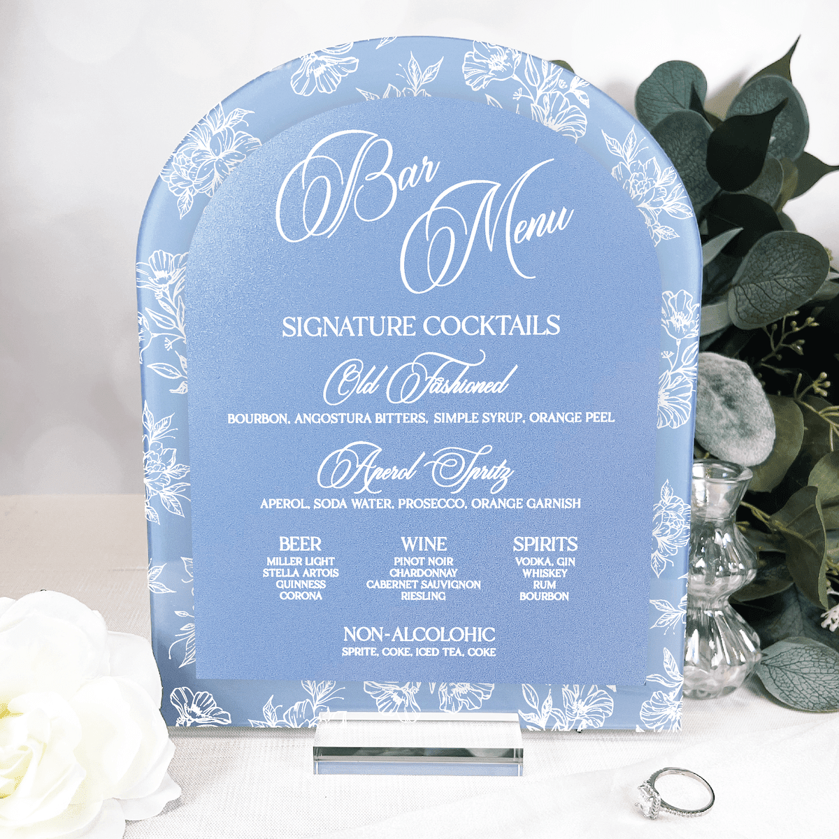 Blue acrylic arch bar menu sign with white floral toile border and script text listing signature cocktails and drink options.