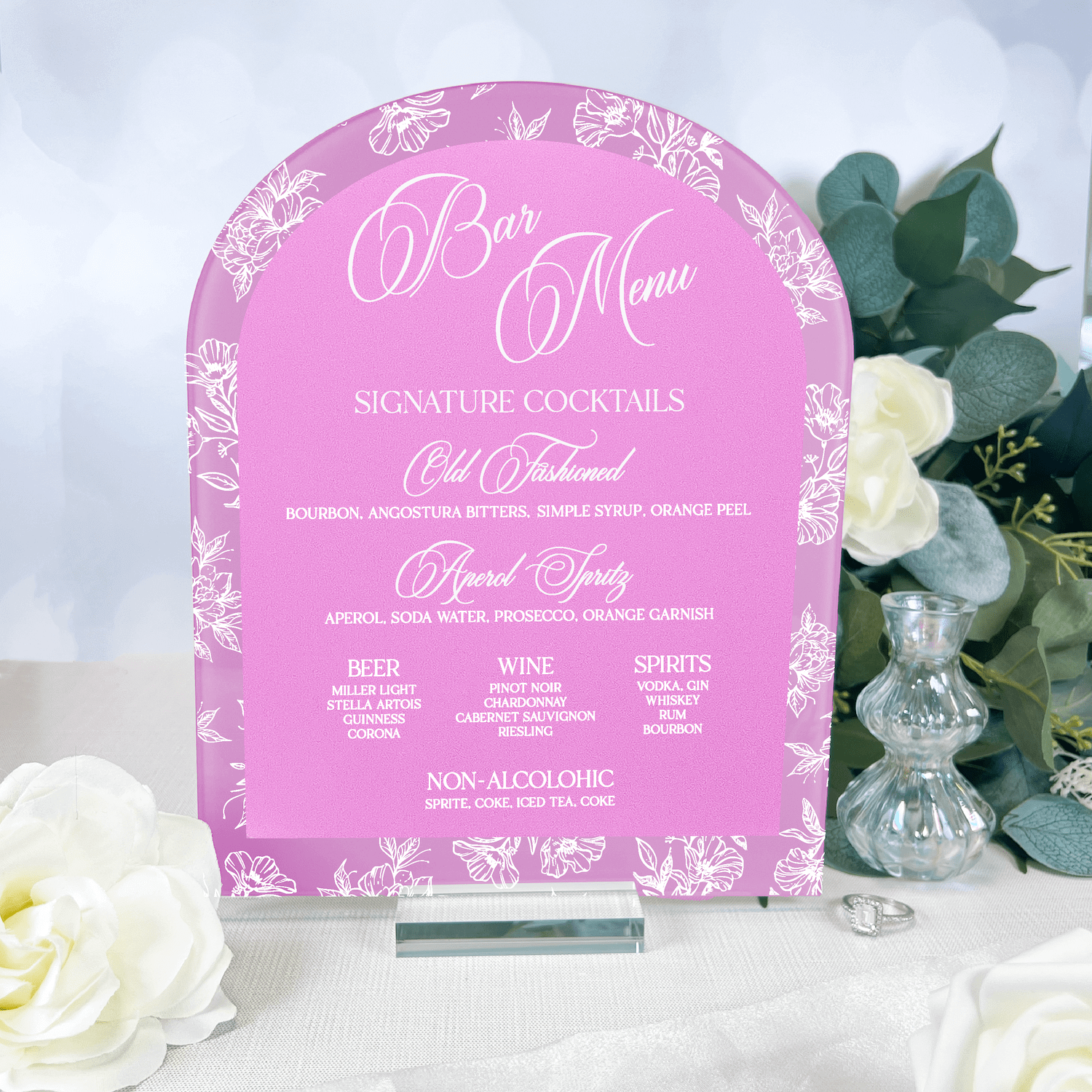 Pink acrylic bar menu sign with white toile floral design and script text, arched top shape, displayed with white roses and greenery.
