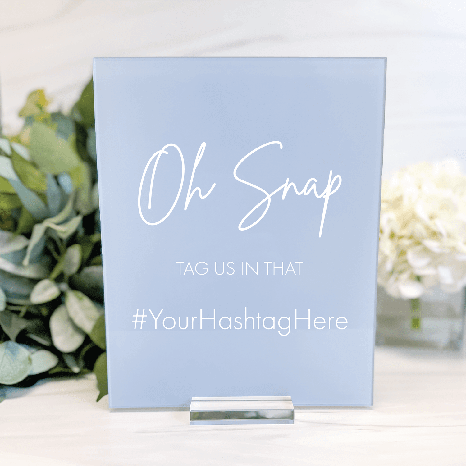 Light blue acrylic sign with white script text reading "Oh Snap" and "TAG US IN THAT #YourHashtagHere" displayed on white surface with greenery.
