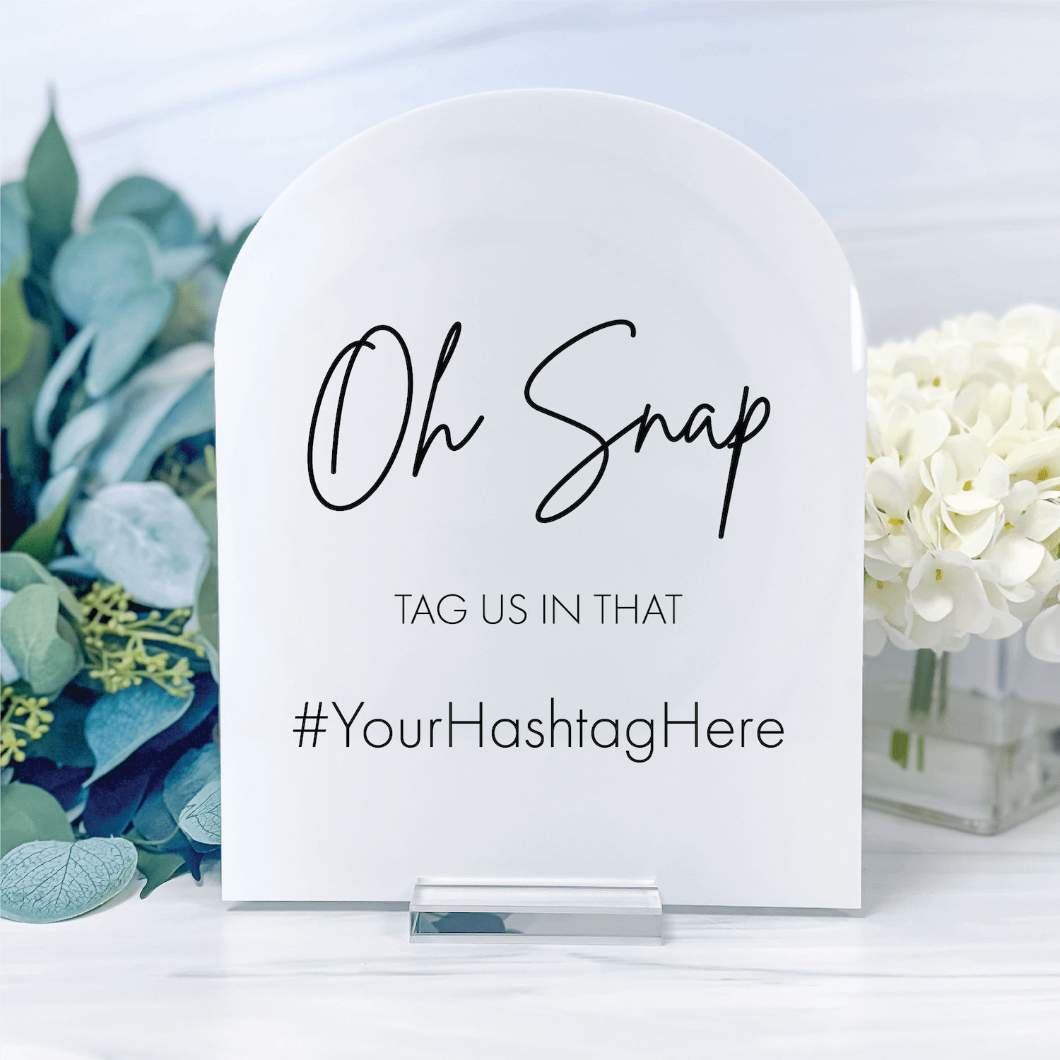 White arched acrylic sign with "Oh Snap" script text, "TAG US IN THAT" and "#YourHashtagHere" in black lettering, displayed with eucalyptus and white flowers.