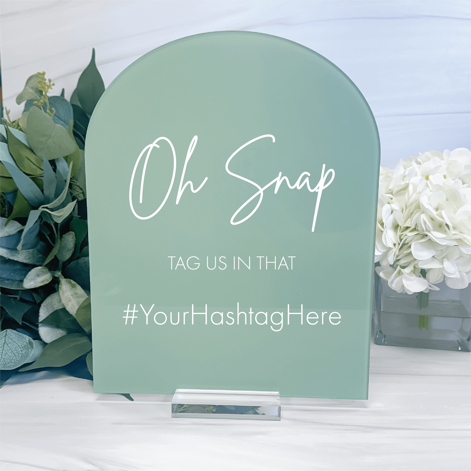 Sage green arched acrylic sign with white script text reading "Oh Snap" and "TAG US IN THAT" with customizable hashtag placeholder, displayed with flowers.