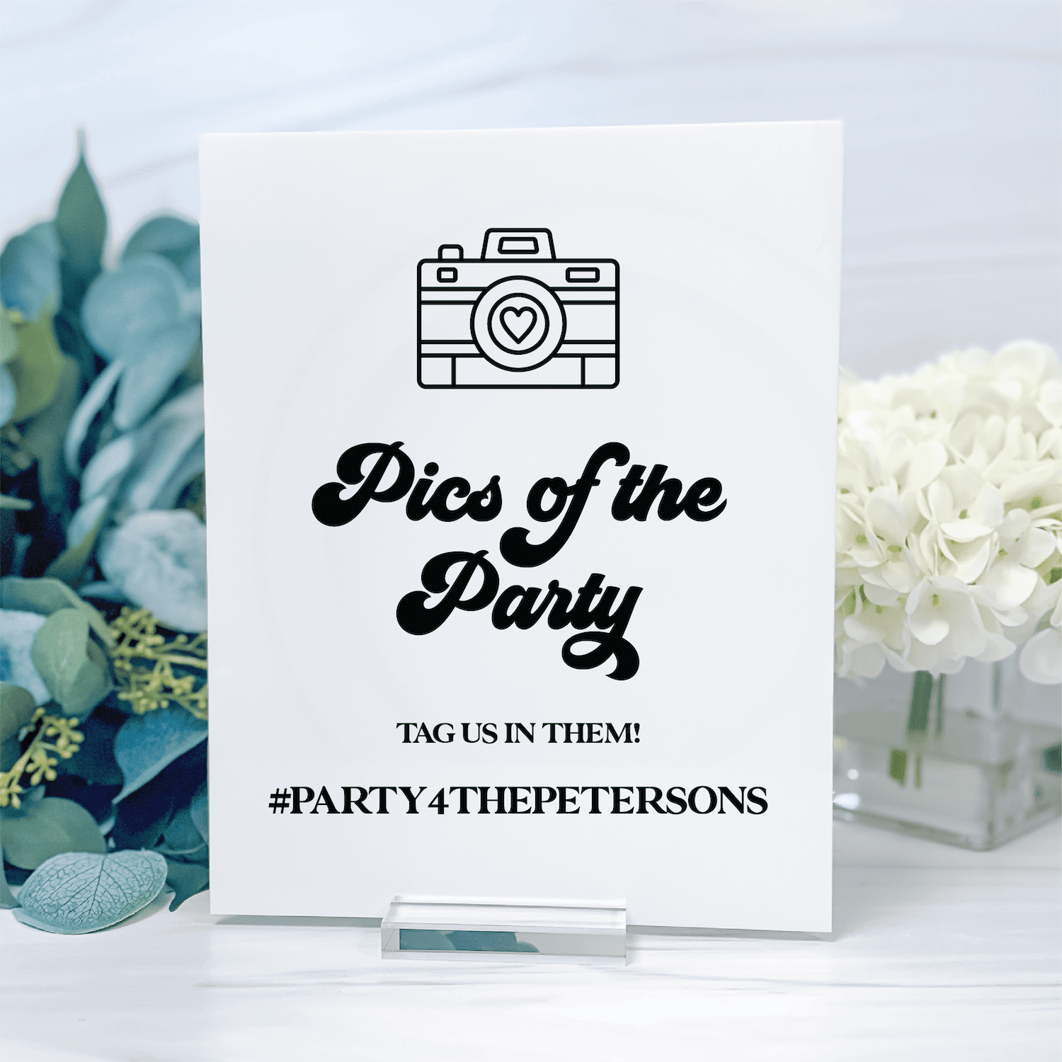 White table sign with camera icon and "Pics of the Party" text, personalized hashtag #PARTY4THEPETERSONS below, flanked by eucalyptus and white flowers.
