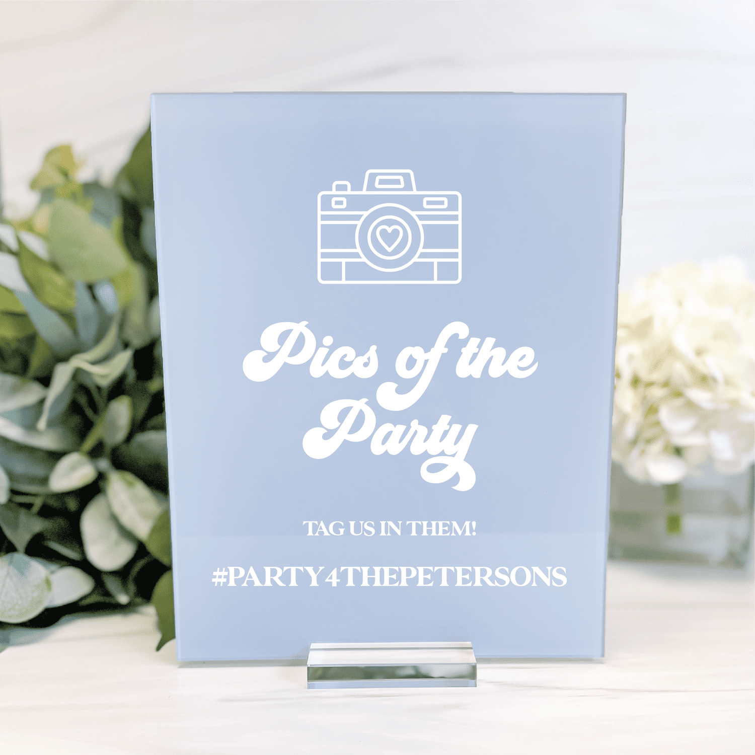 Light blue acrylic sign with white camera icon and "Pics of the Party" text, hashtag #PARTY4THEPETERSONS below, displayed with flowers and greenery.