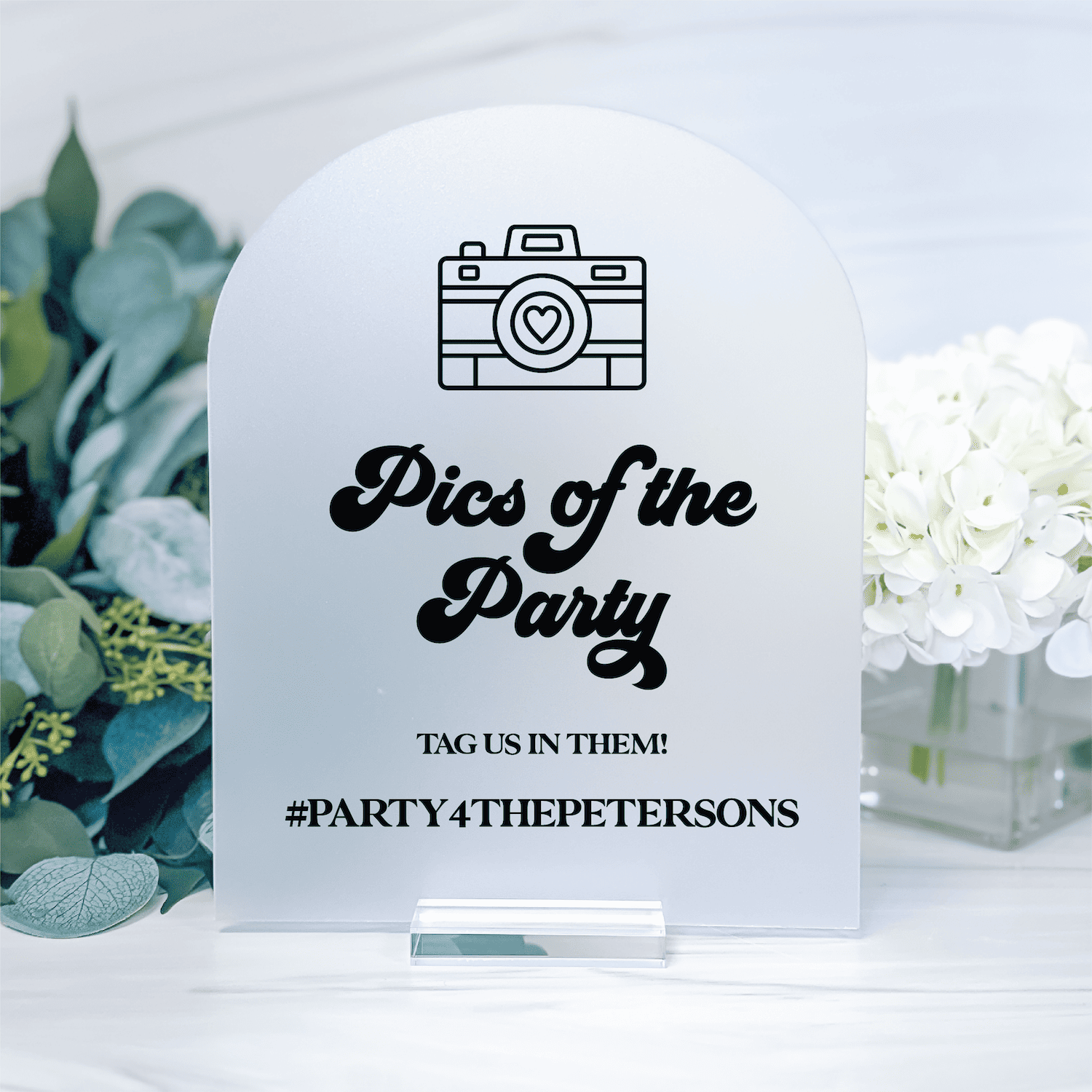 White acrylic table sign with camera icon, "Pics of the Party" script text, and personalized hashtag #PARTY4THEPETERSONS for wedding photo display.