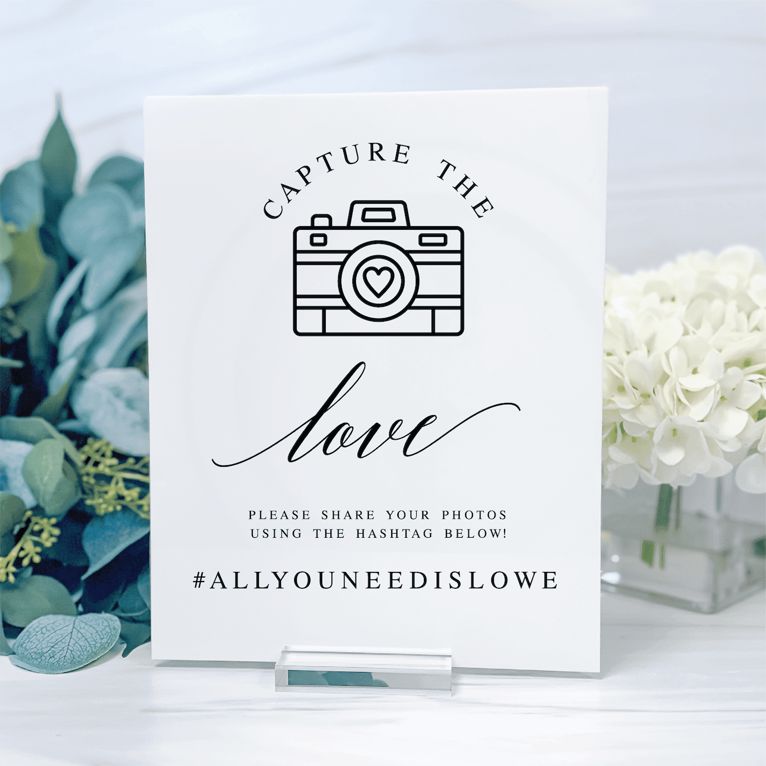White wedding sign with camera icon, "Capture the Love" text, and hashtag #ALLYOUNEEDISLOVE, displayed with eucalyptus and white flowers.