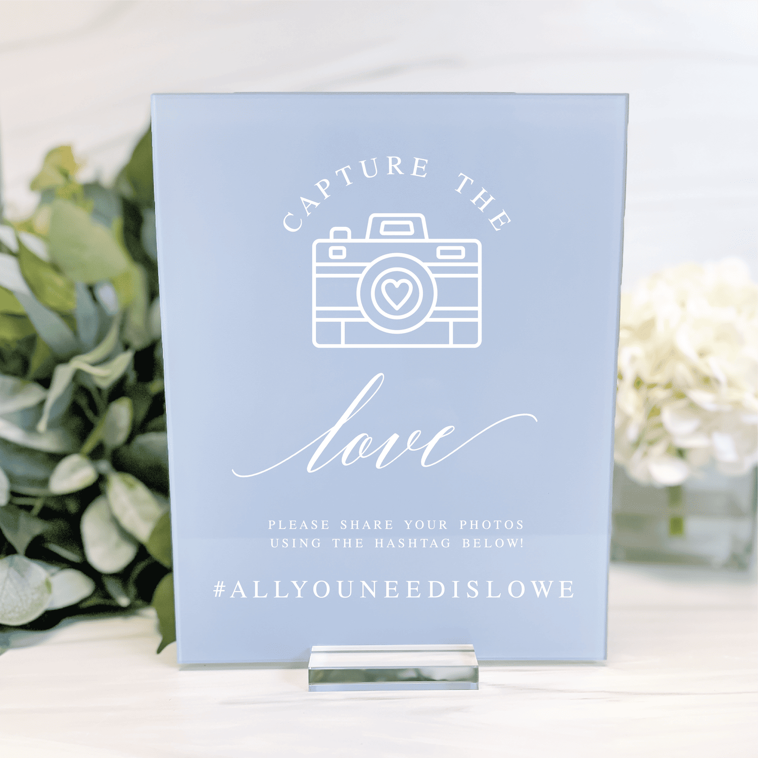 Light blue acrylic sign with white camera icon, "Capture the Love" text, and hashtag #allyouneedislove wedding design