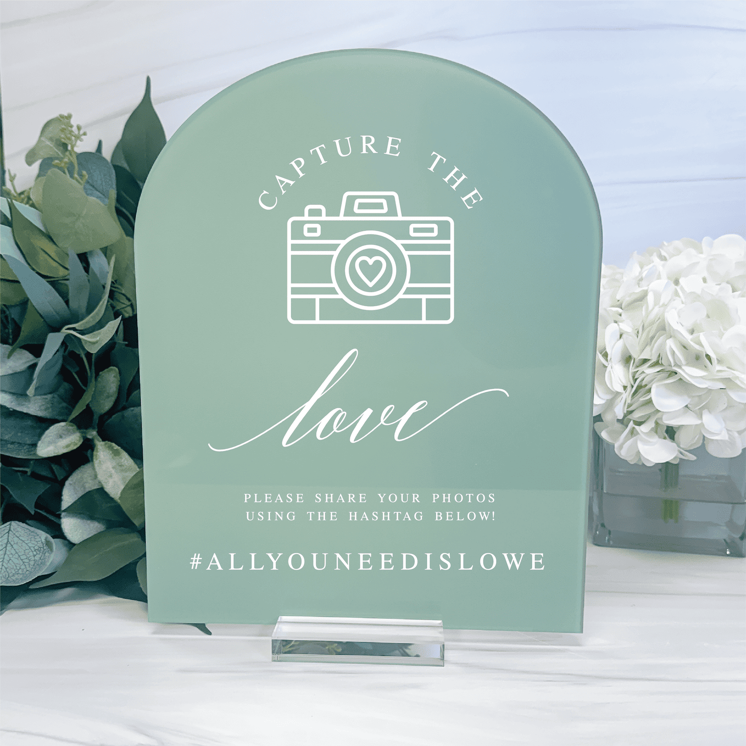 Sage green arch-shaped acrylic wedding sign with camera icon, "love" script, and hashtag text for photo sharing displays