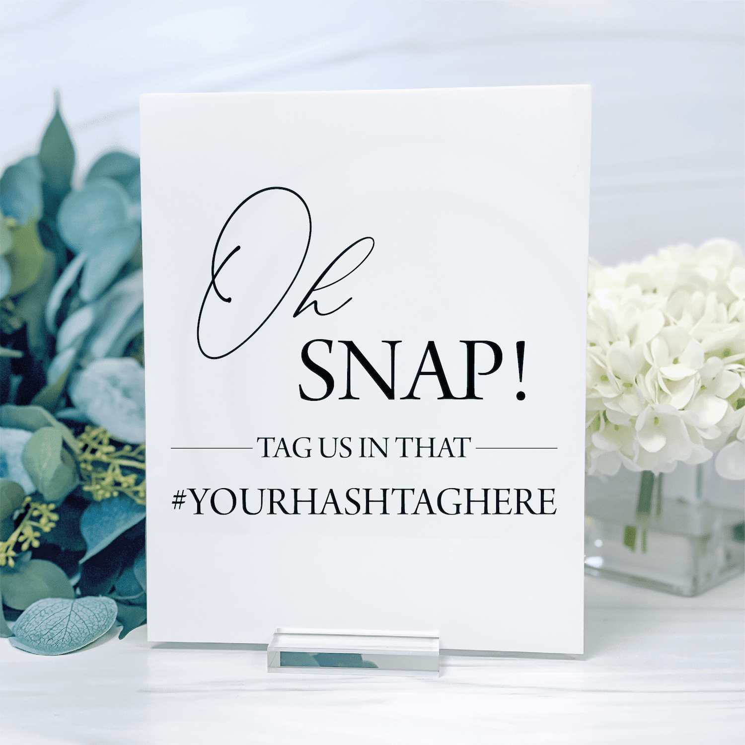 White table sign with "Oh SNAP!" text and hashtag prompt, displayed with eucalyptus and white flowers