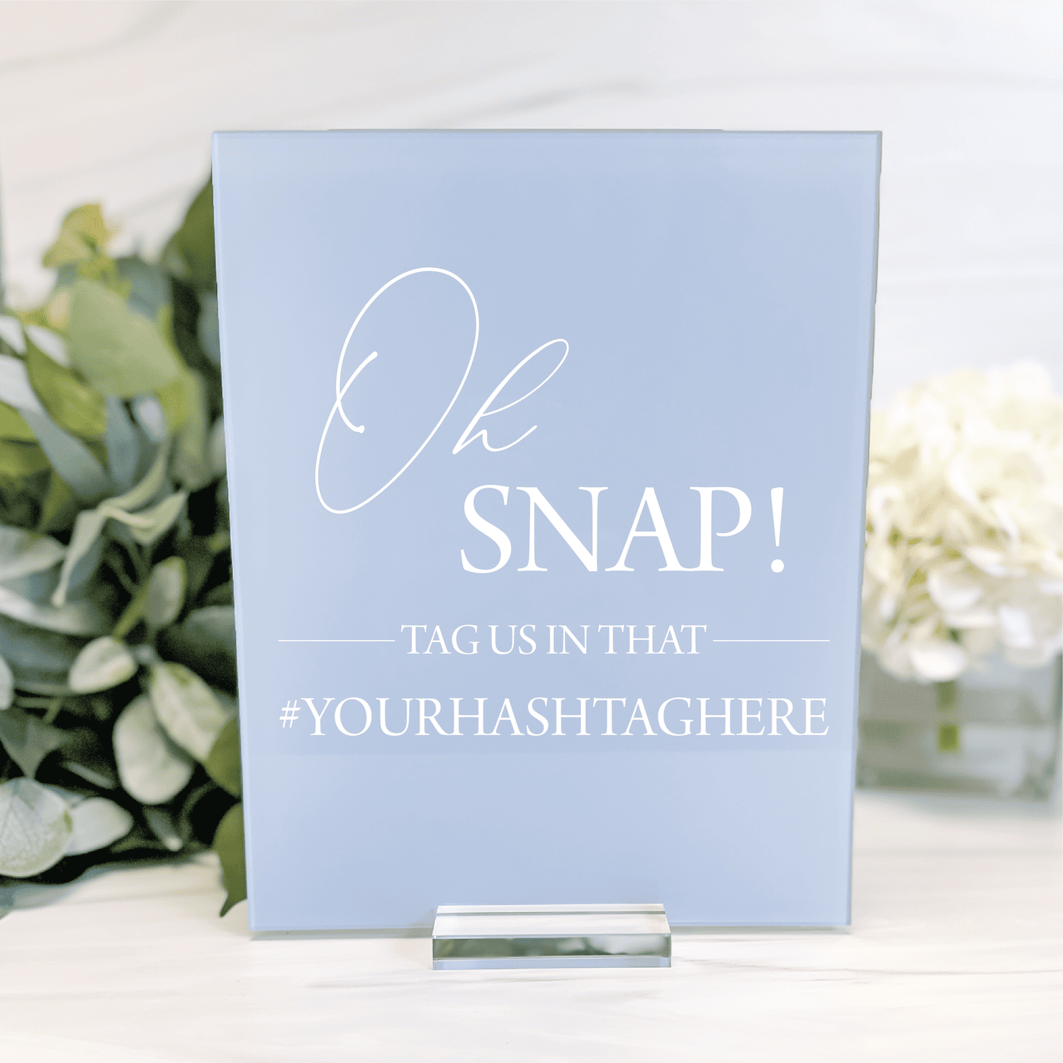 Light blue acrylic sign with white script text reading "Oh SNAP! Tag us in that #yourhashtghere" displayed on stand with greenery.