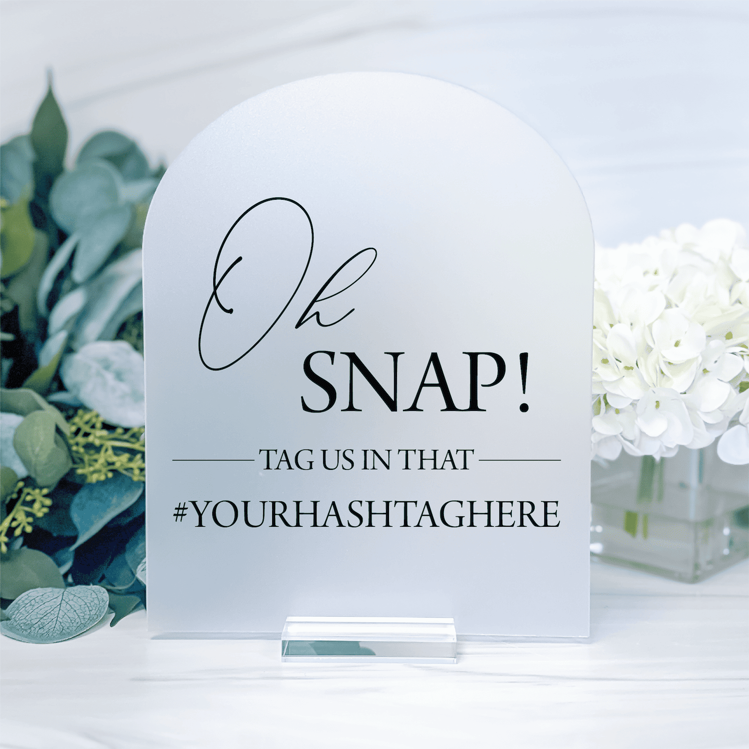 Frosted acrylic wedding sign with "Oh SNAP!" text and hashtag prompt, displayed with eucalyptus and white flowers.