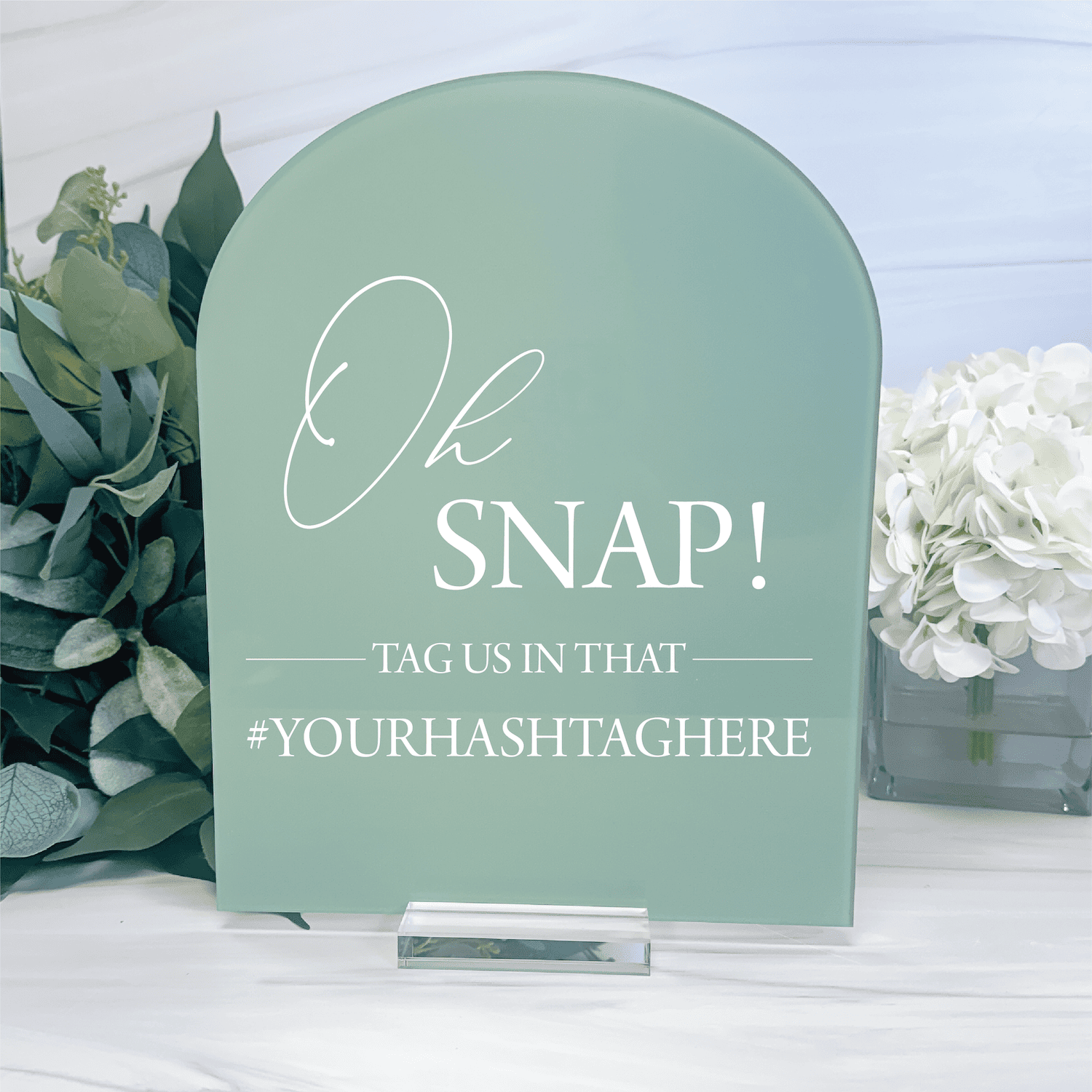 Sage green arch-shaped acrylic sign with "Oh SNAP! Tag Us In That #YourHashtagHere" text in white lettering, displayed with eucalyptus and white flowers.