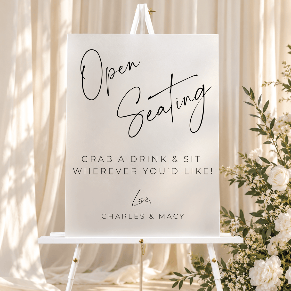 Modern rectangular open seating wedding sign with black script lettering, displayed on a white easel with ivory drapery and flowers.