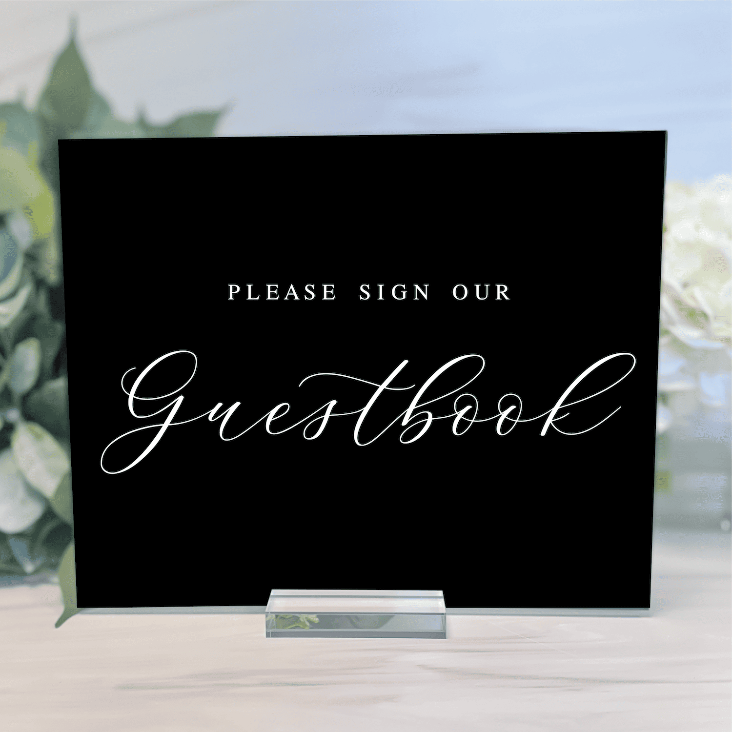 Black guestbook sign with "Please sign our Guestbook" in white cursive text, displayed on small stand with greenery background.