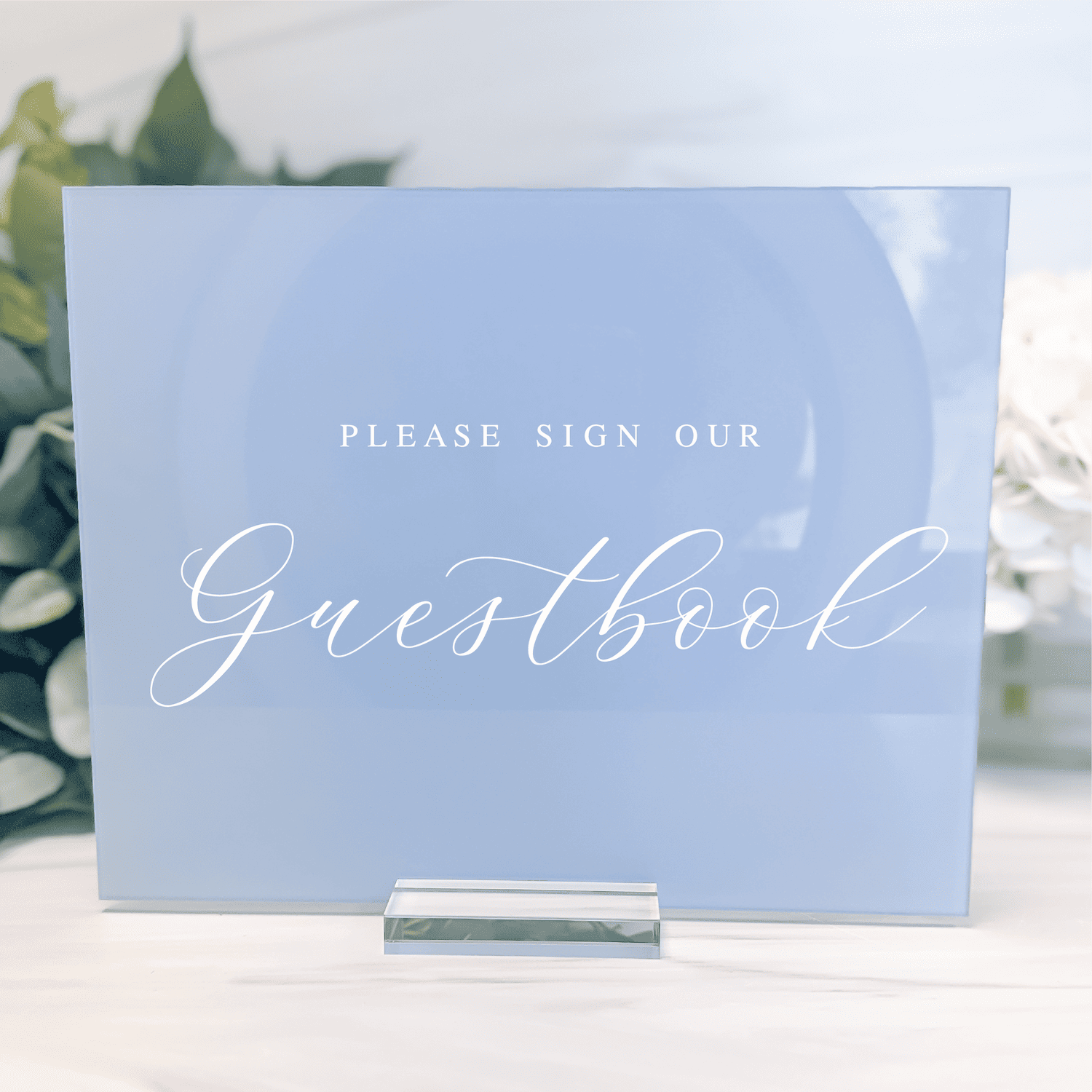 Light blue acrylic guestbook sign with "Please Sign Our Guestbook" text in white script and sans-serif fonts, displayed on a clear stand.