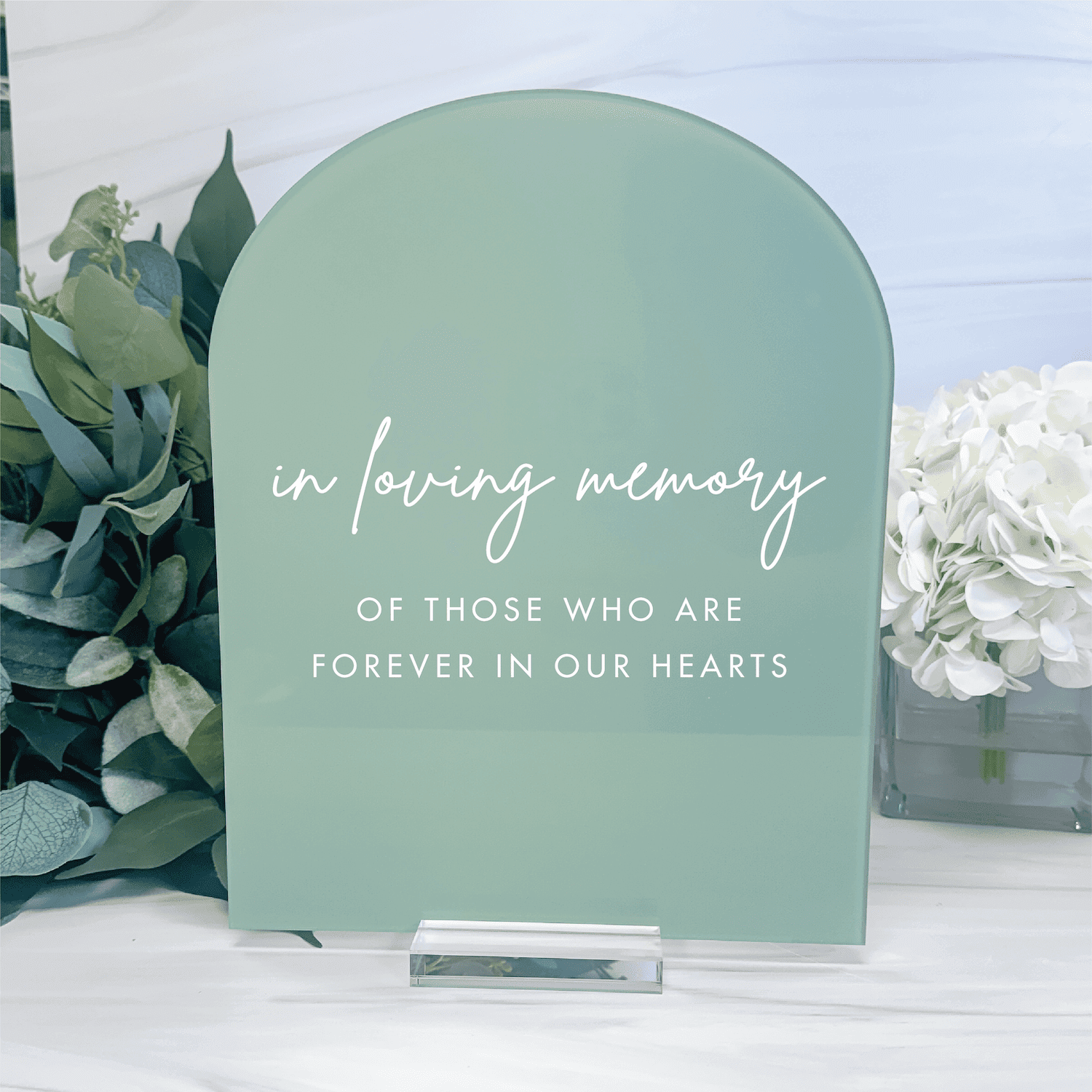 Sage green arched acrylic sign with "in loving memory of those who are forever in our hearts" text, displayed with white flowers.