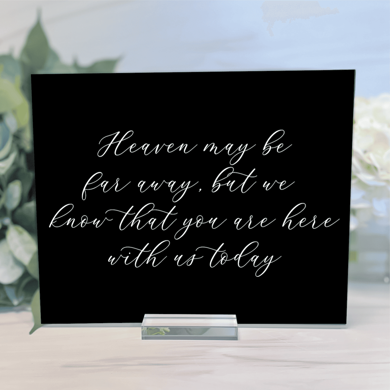 Black acrylic memorial sign with white script text reading "Heaven may be far away, but we know that you are here with us today" displayed on stand with floral backdrop