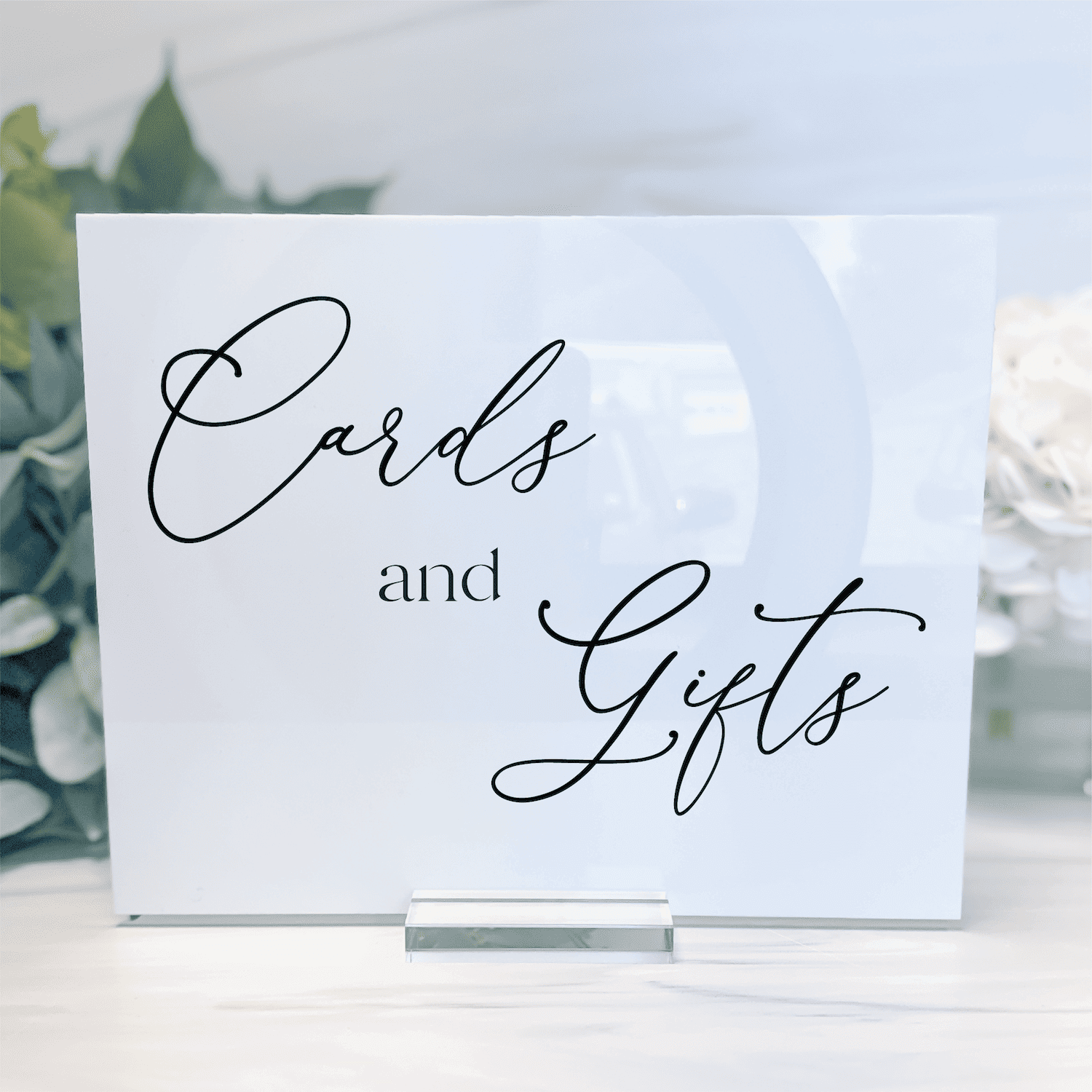 White acrylic wedding sign with "Cards and Gifts" in elegant black script, displayed on a clear stand with greenery in background.