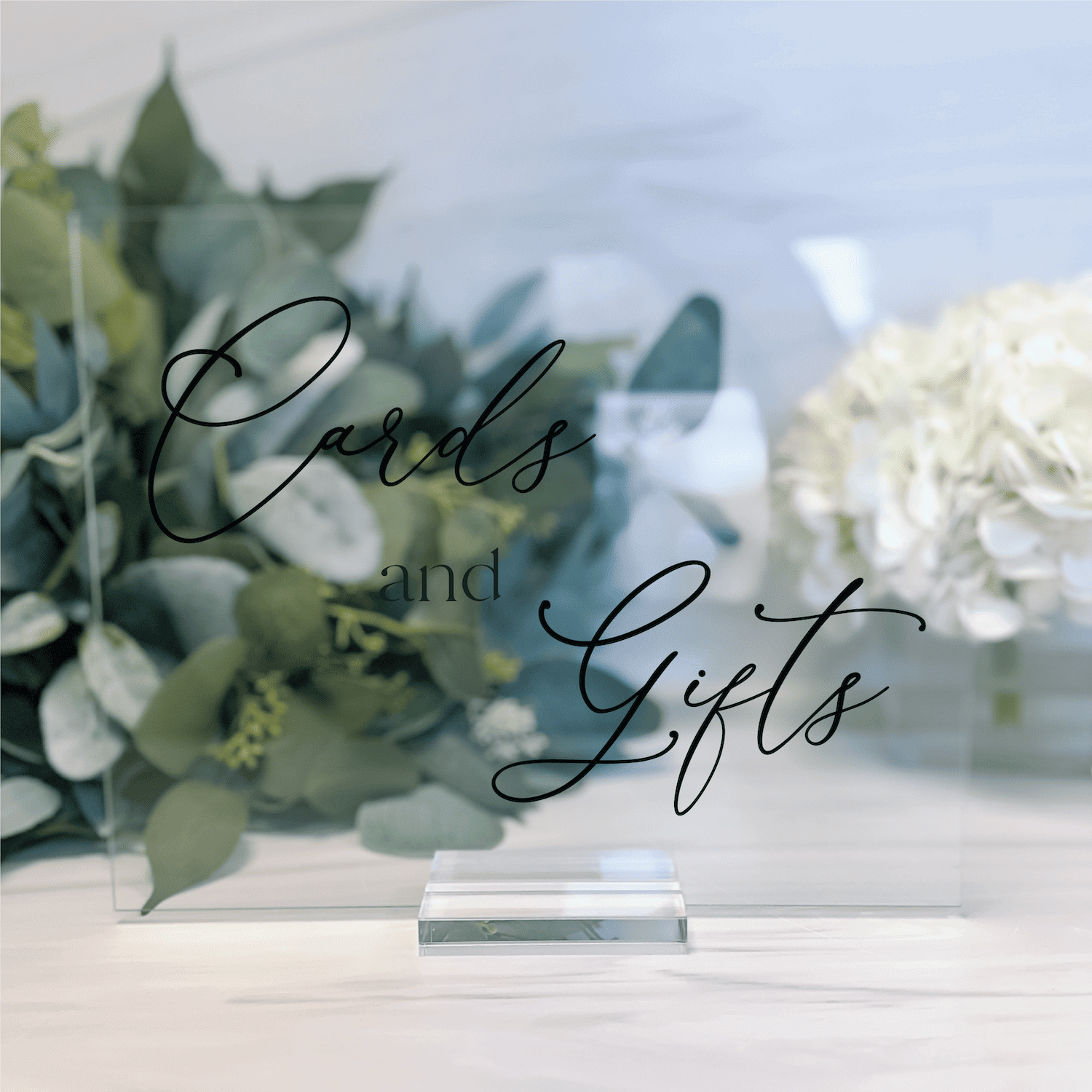 Clear acrylic wedding sign with "Cards and Gifts" in elegant script, displayed on table with eucalyptus greenery and white flowers.