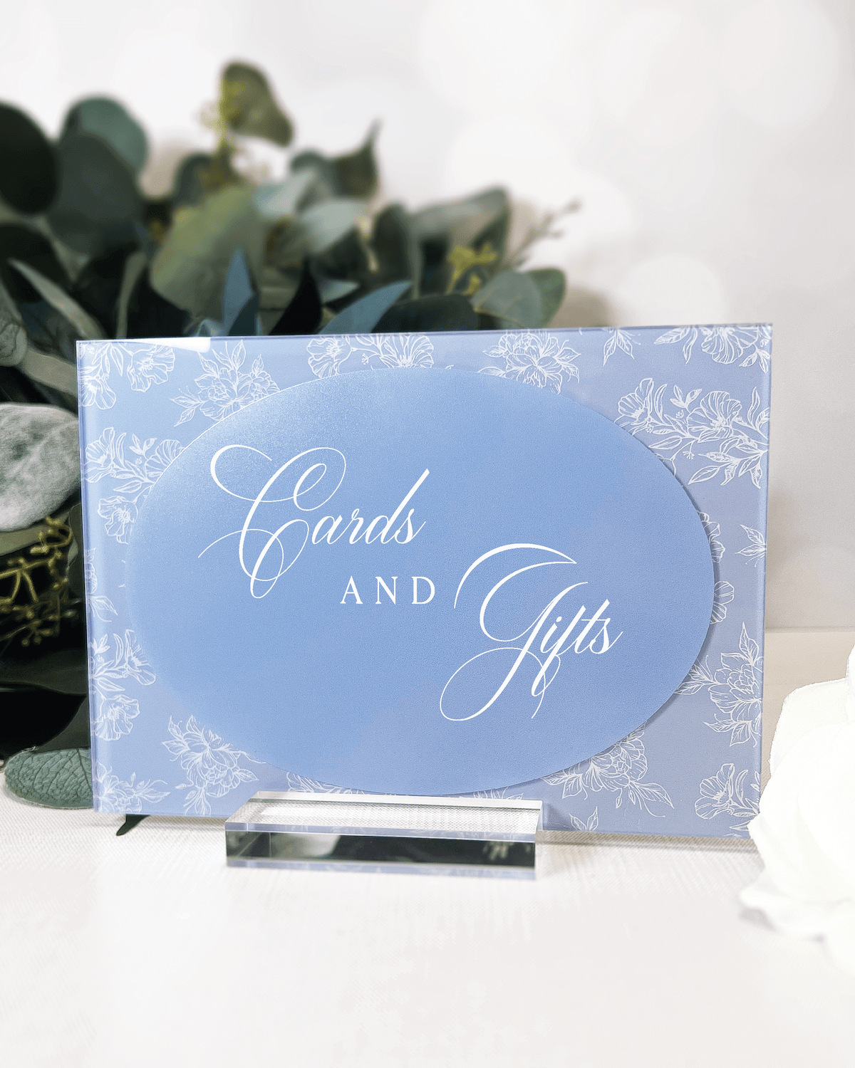 Periwinkle acrylic sign with white floral toile border and cursive text reading "Cards and Gifts" displayed on a stand with eucalyptus backdrop.