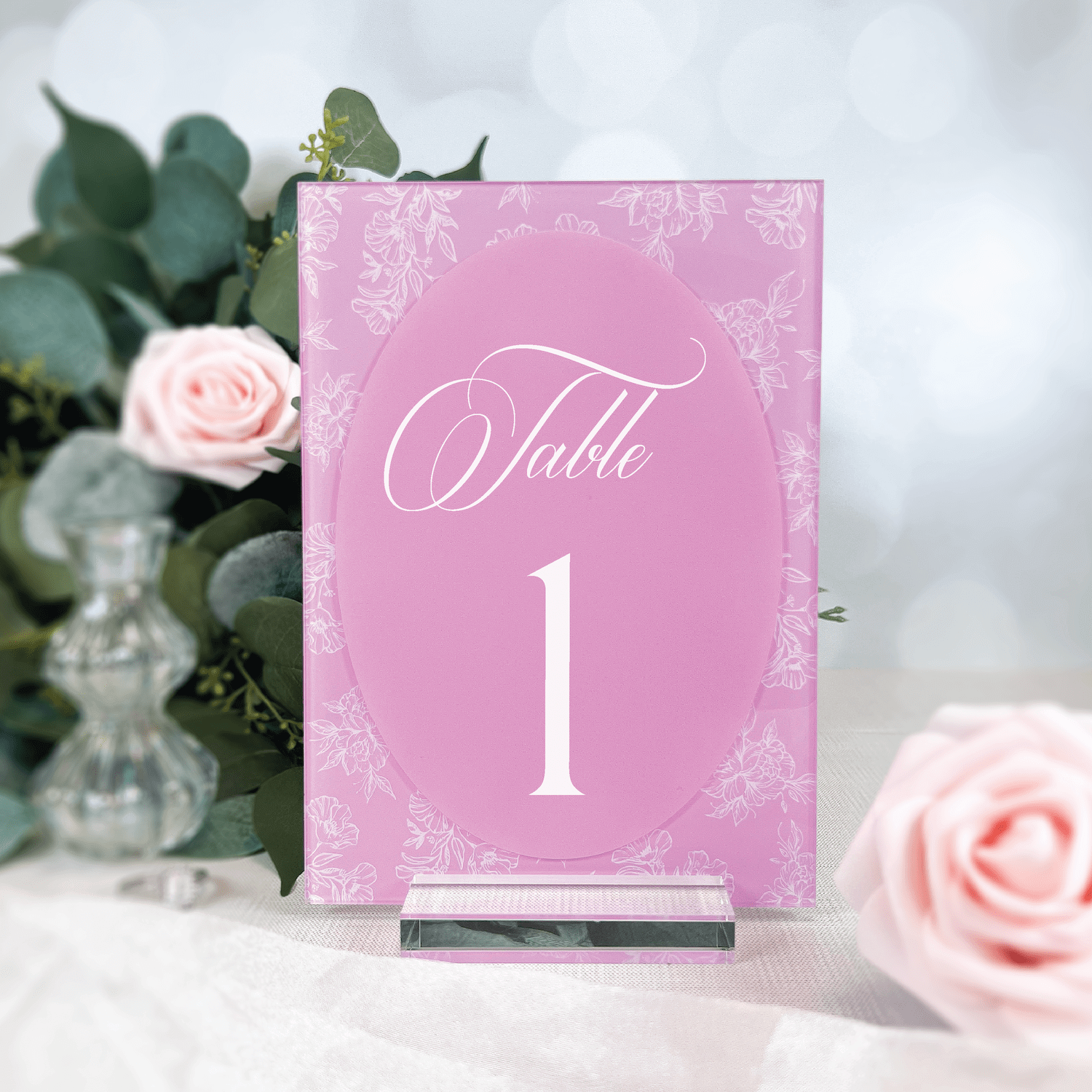 Pink acrylic table number card with white "Table 1" text and floral chinoiserie border design, displayed with roses and greenery.