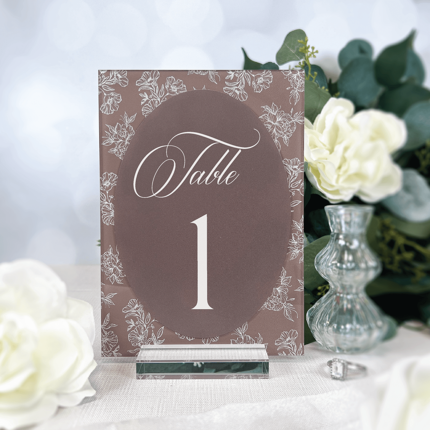 Taupe acrylic table number card with ornate floral border design, displaying "Table 1" in elegant script and numerals, surrounded by white roses and greenery.