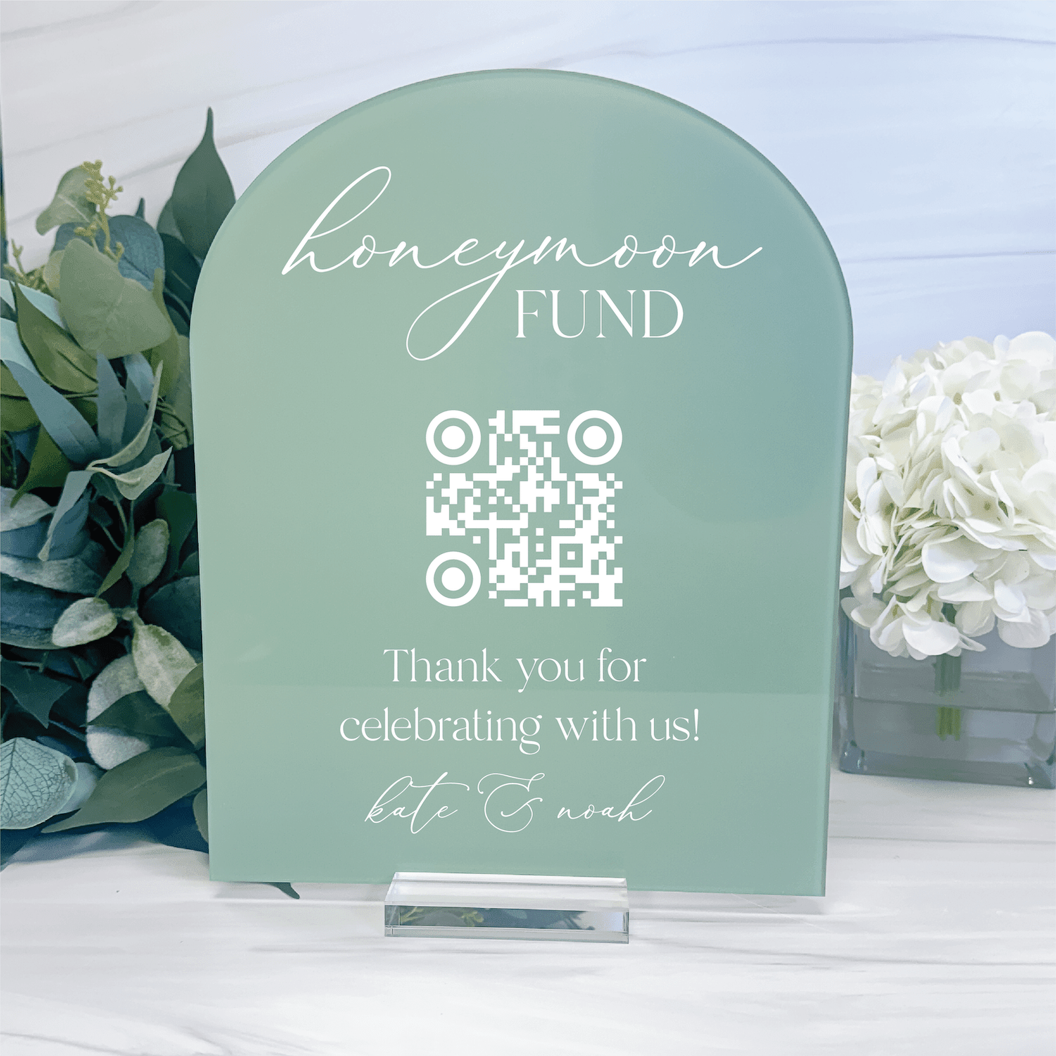 Sage green arched acrylic sign with "honeymoon FUND" text, QR code, and personalized names at bottom, displayed with flowers