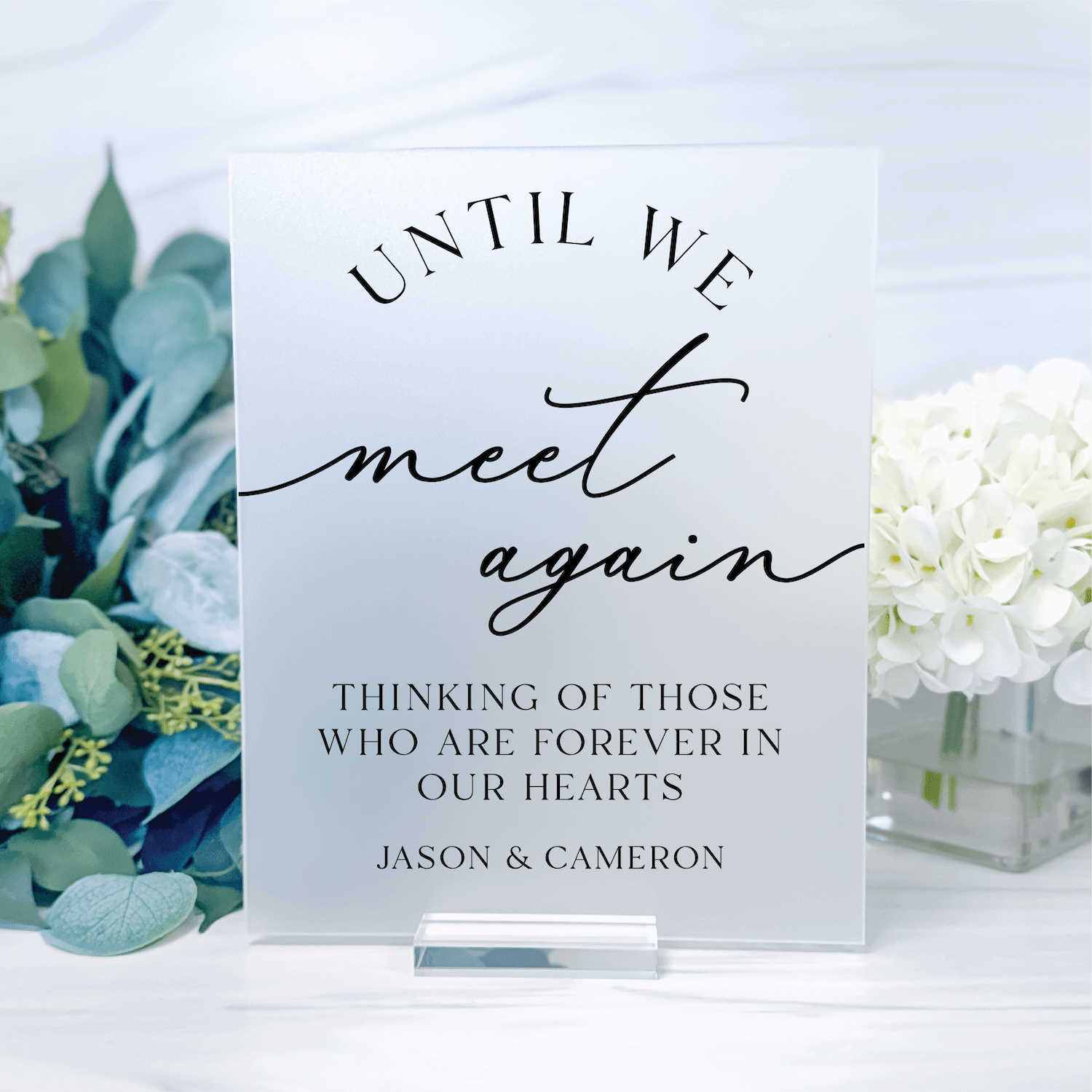 White acrylic sign with "Until We Meet Again" text and personalization names Jason & Cameron, displayed with white flowers and eucalyptus.