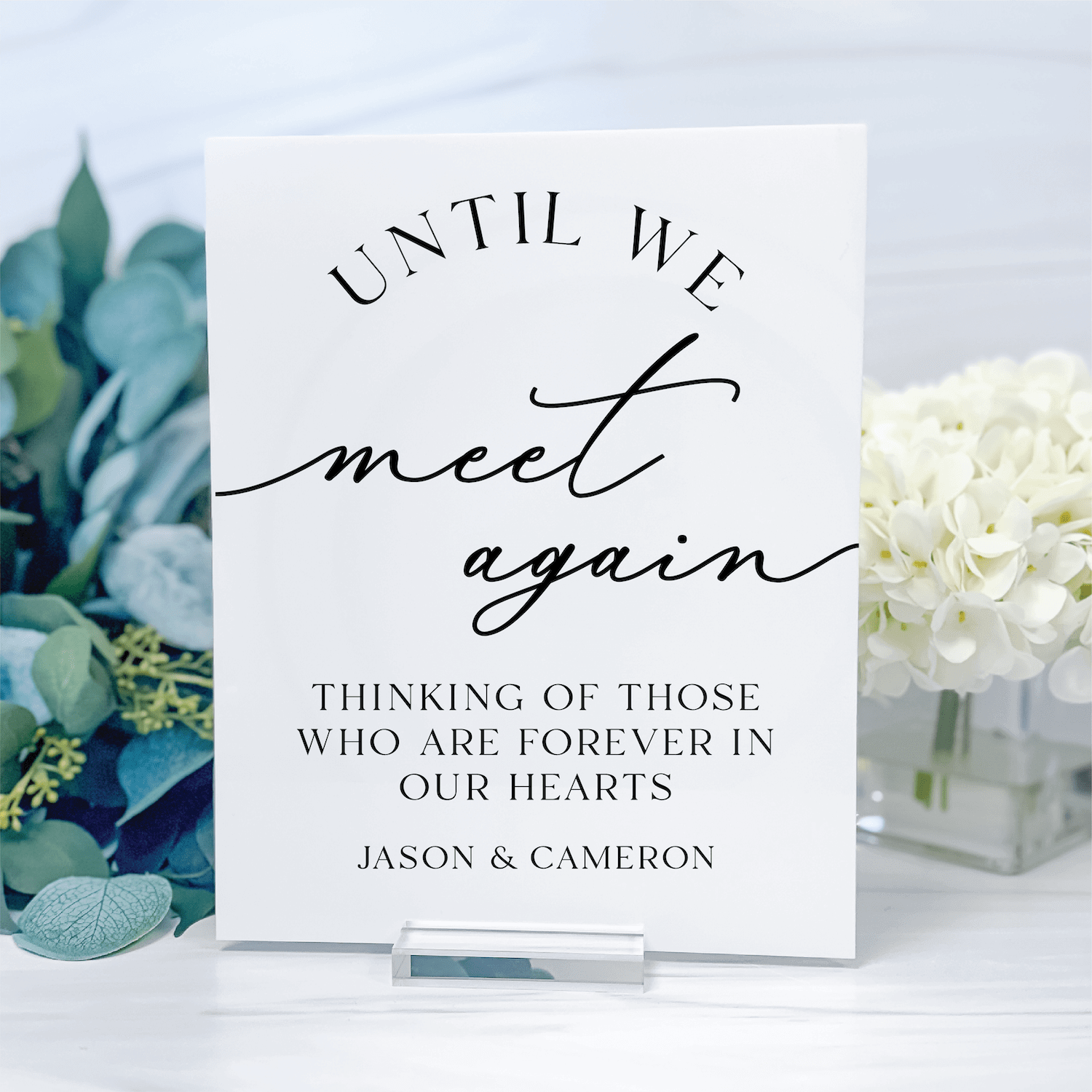 White acrylic sign with black cursive and serif text reading "Until We Meet Again" and "Thinking Of Those Who Are Forever In Our Hearts" with names Jason & Cameron, displayed with eucalyptus and white flowers.