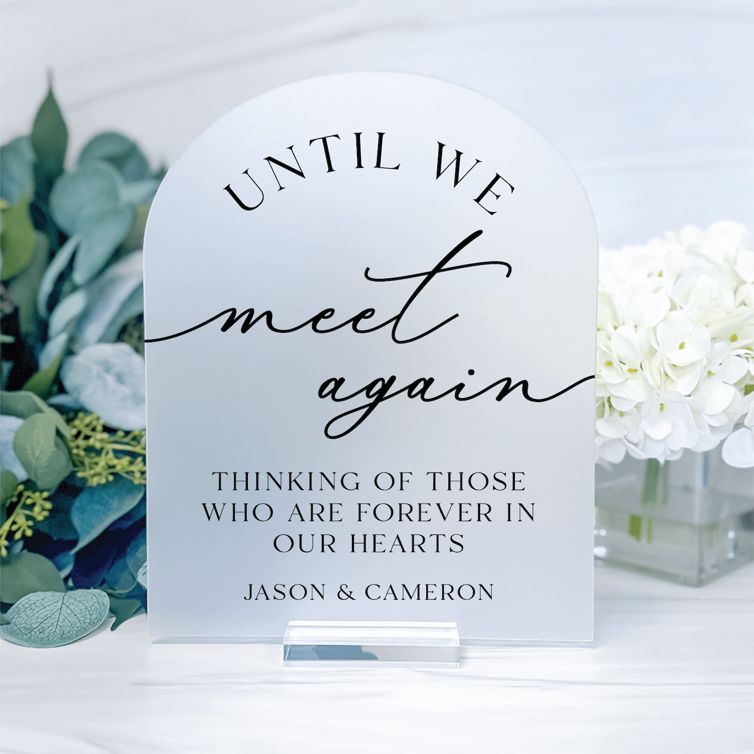 White acrylic arch sign with "Until We Meet Again" in script and serif fonts, personalized with names Jason & Cameron, surrounded by white flowers and greenery.