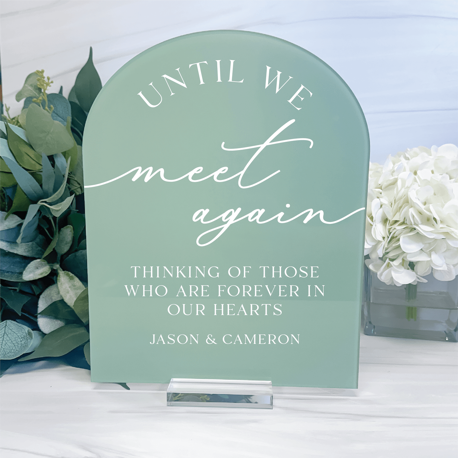 Sage green arched acrylic sign with white script text reading "Until We Meet Again" and personalized names Jason & Cameron, memorial table sign with flowers.
