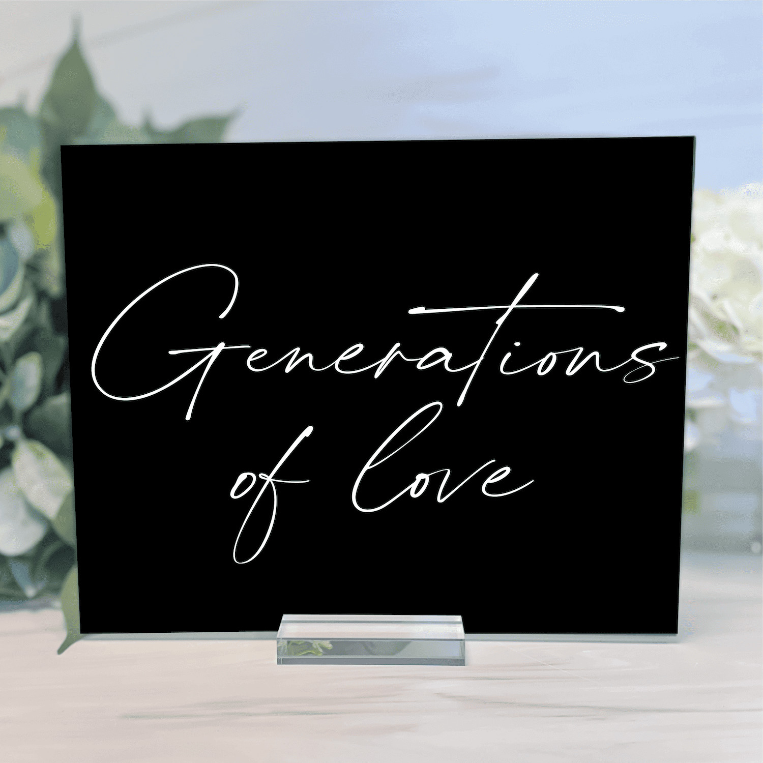 Black acrylic wedding sign with "Generations of Love" in elegant white script lettering, displayed on a silver stand with greenery in background.