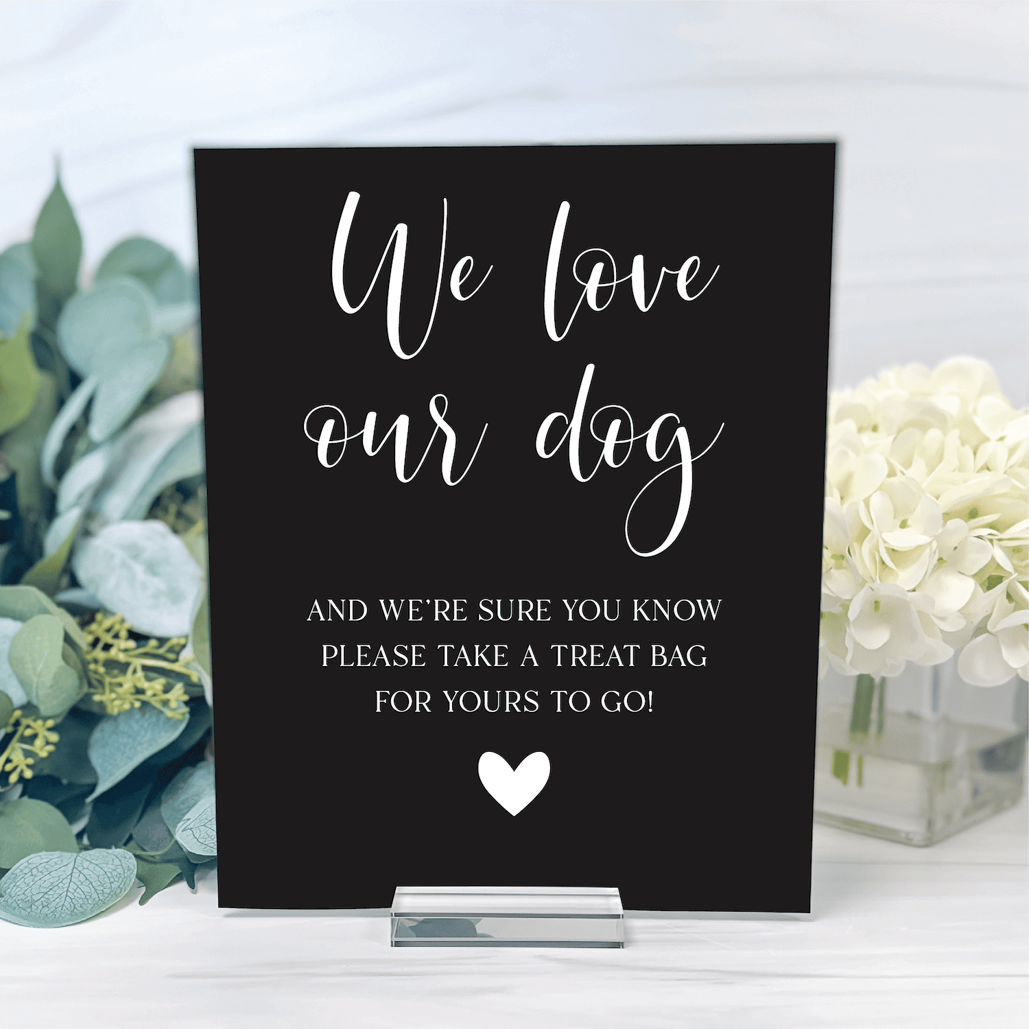 Black acrylic sign with white script text reading "We love our dog" and treat bag message, displayed with white flowers and greenery