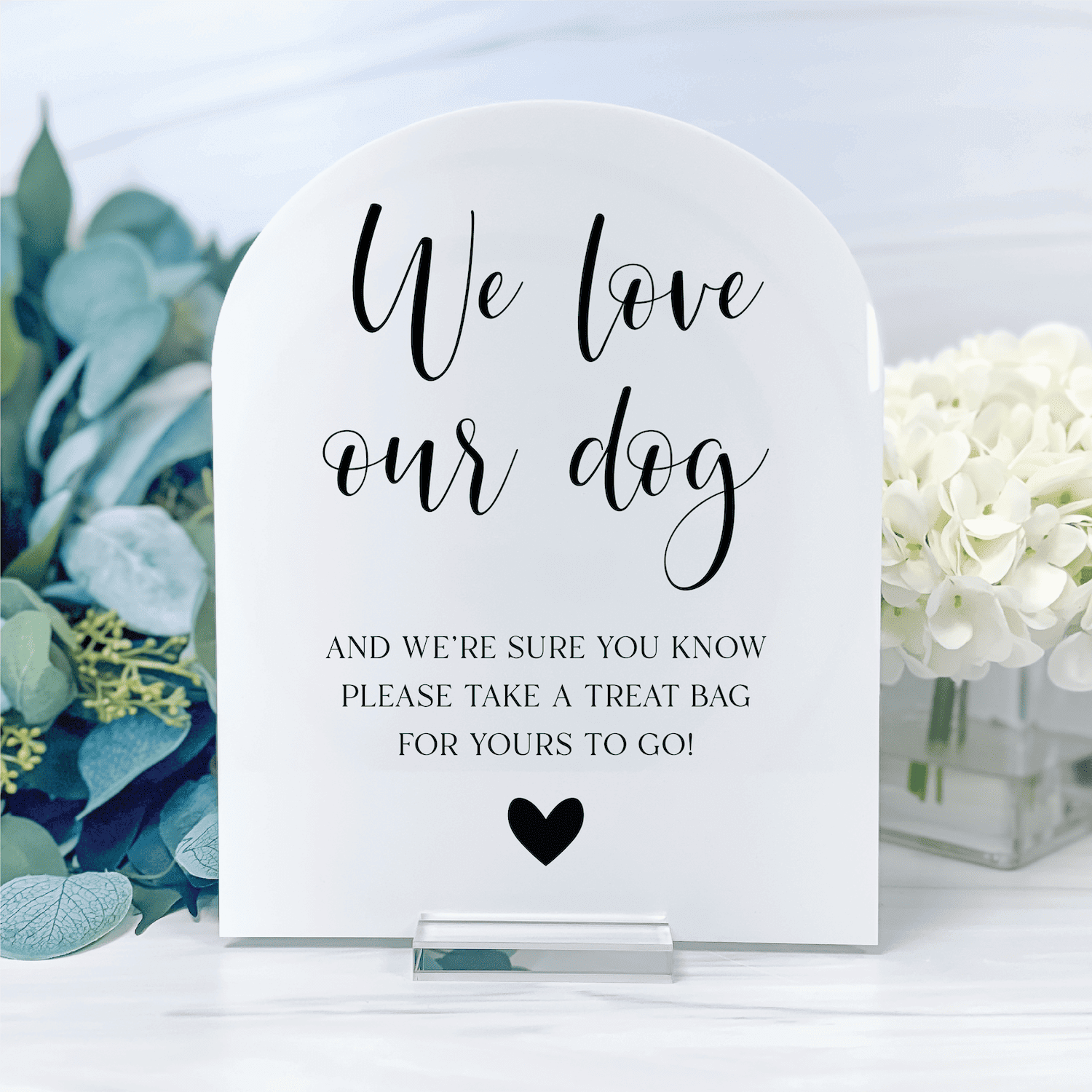 White arched acrylic sign with black script text reading "We love our dog" and smaller text below about treat bags, with a heart symbol, displayed on a table with white flowers and greenery.