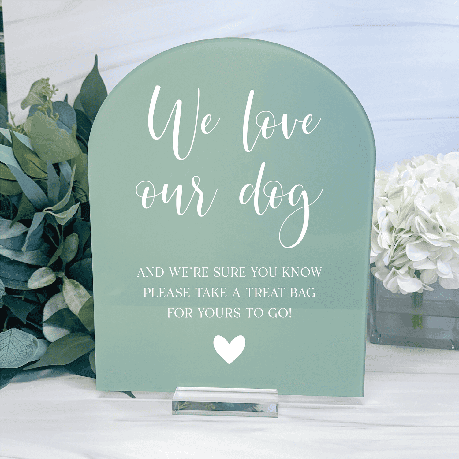 Sage green acrylic arch sign with white script text reading "We love our dog" and smaller text about treat bags, decorated with a heart, flanked by greenery and white flowers