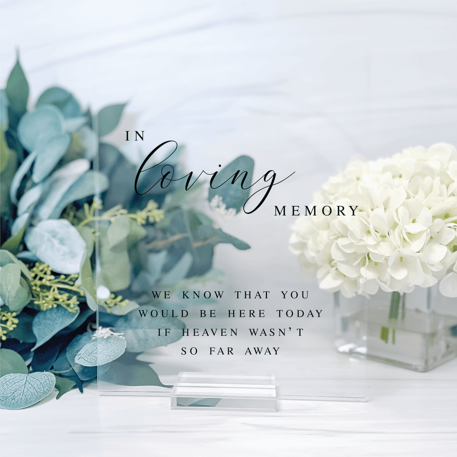 Clear acrylic sign with "In Loving Memory" text and sympathy message, displayed with white hydrangeas and eucalyptus flowers.